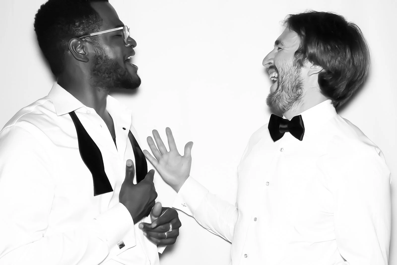 Two men dressed in white shirts and black bow ties laughing and engaging in a lively conversation against a plain white background.