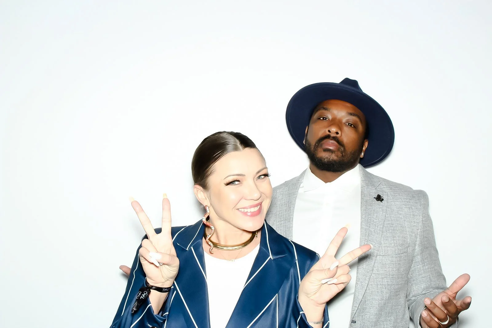 Two stylish people, a woman and a man, posing together against a plain white background. The woman is smiling and making peace signs with both hands; she has brown hair, wears earrings and a dark blazer with white lines. The man wears a gray suit, white shirt, and a dark wide-brimmed hat, with a serious expression.