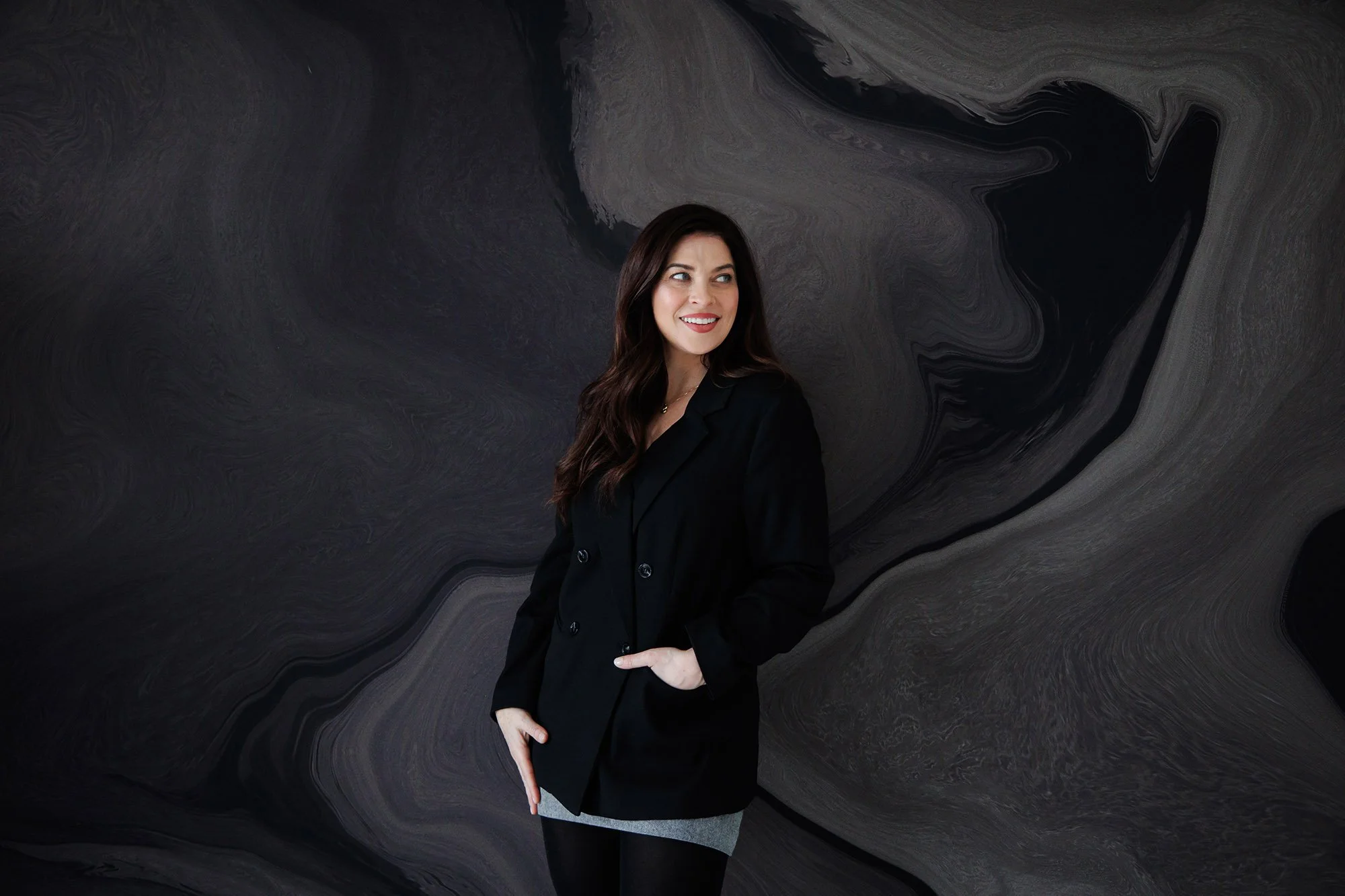 A woman with long dark hair, wearing a black blazer and grey skirt, standing against a black, grey, and white marbled background, smiling and looking to her right.