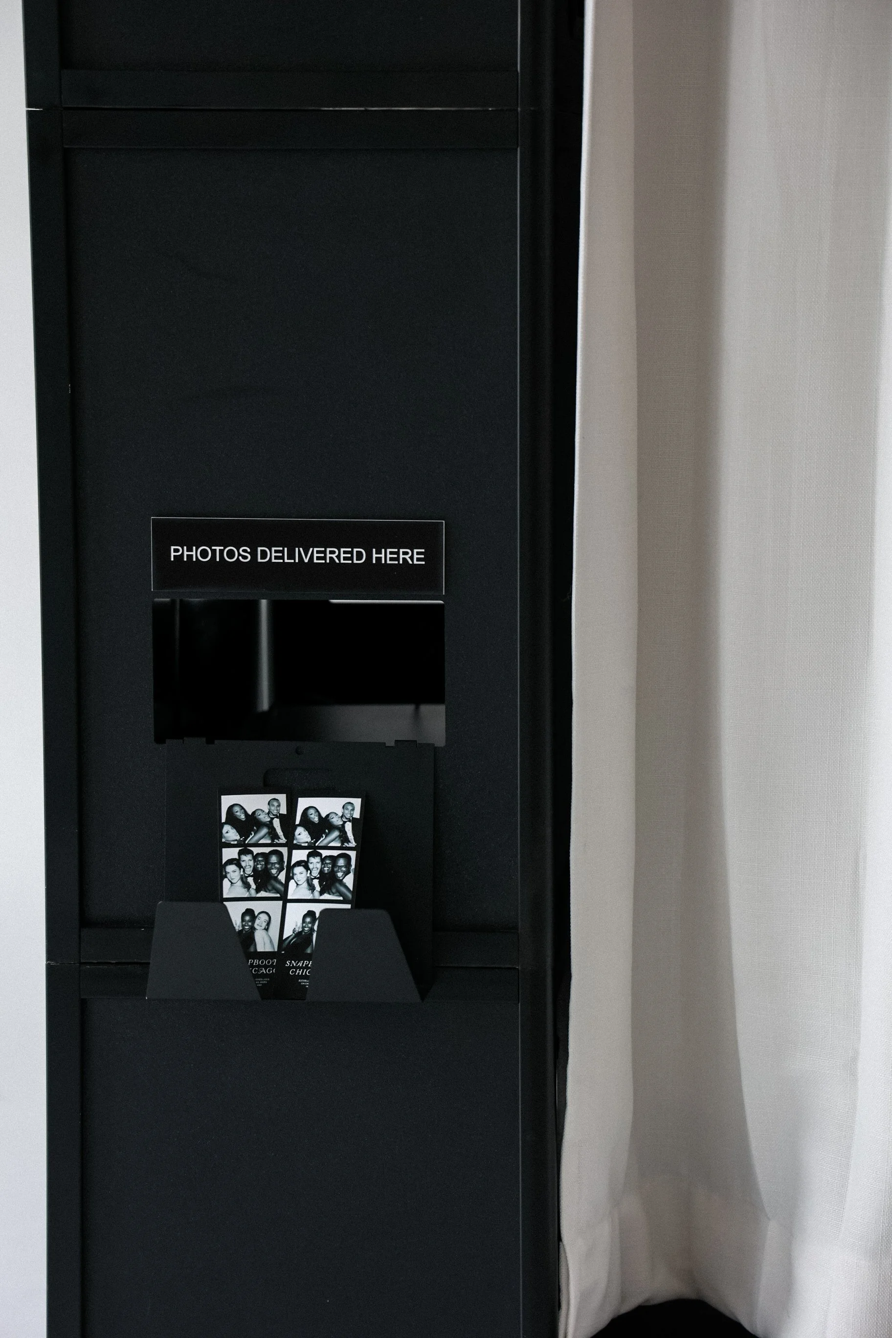 Photo booth with a sign that says 'Photos Delivered Here' and black-and-white photo strips displayed.