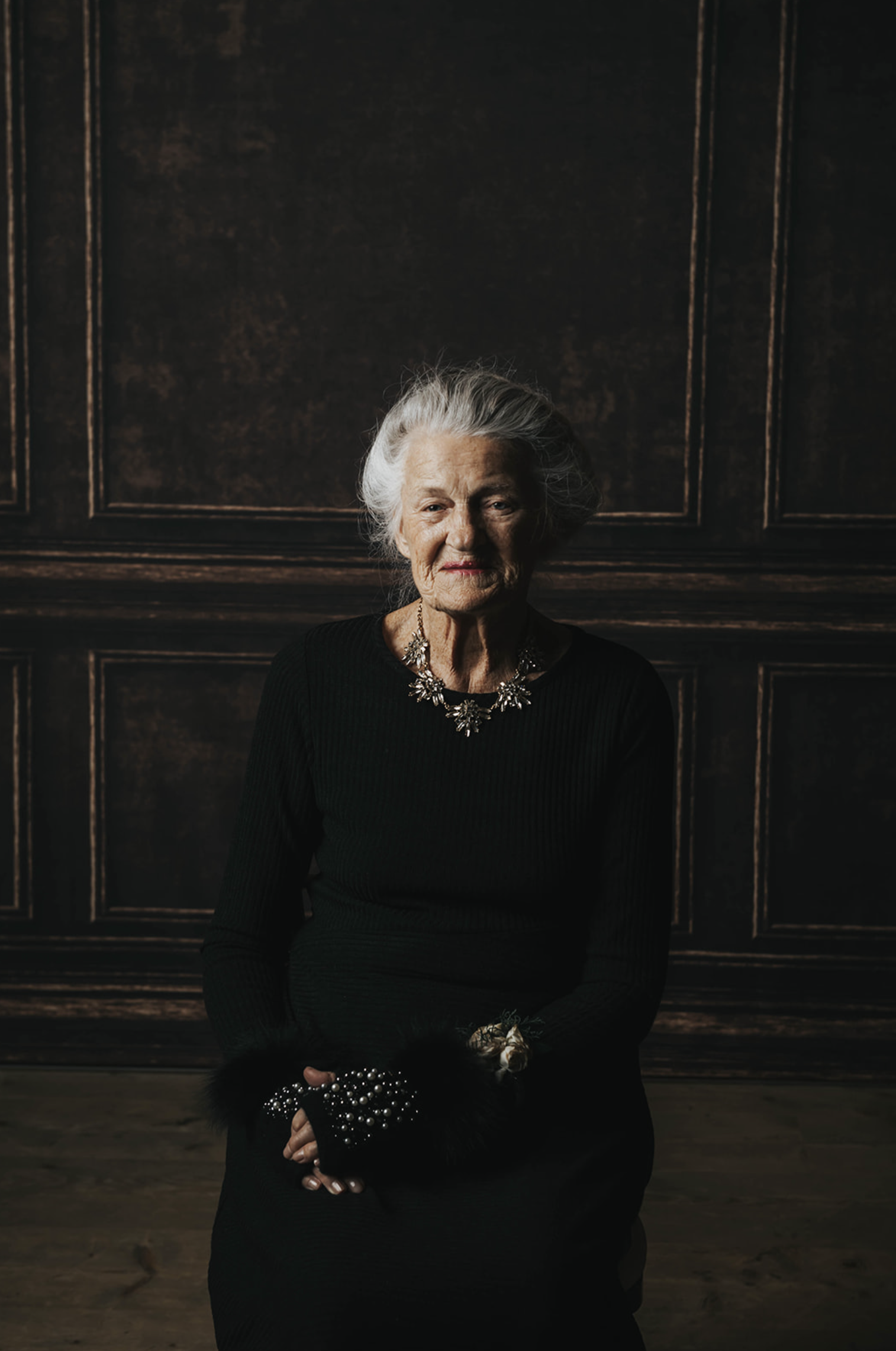 An elderly woman with white hair wearing a black dress and pearl accessories, sitting in front of a dark brown paneled wall.