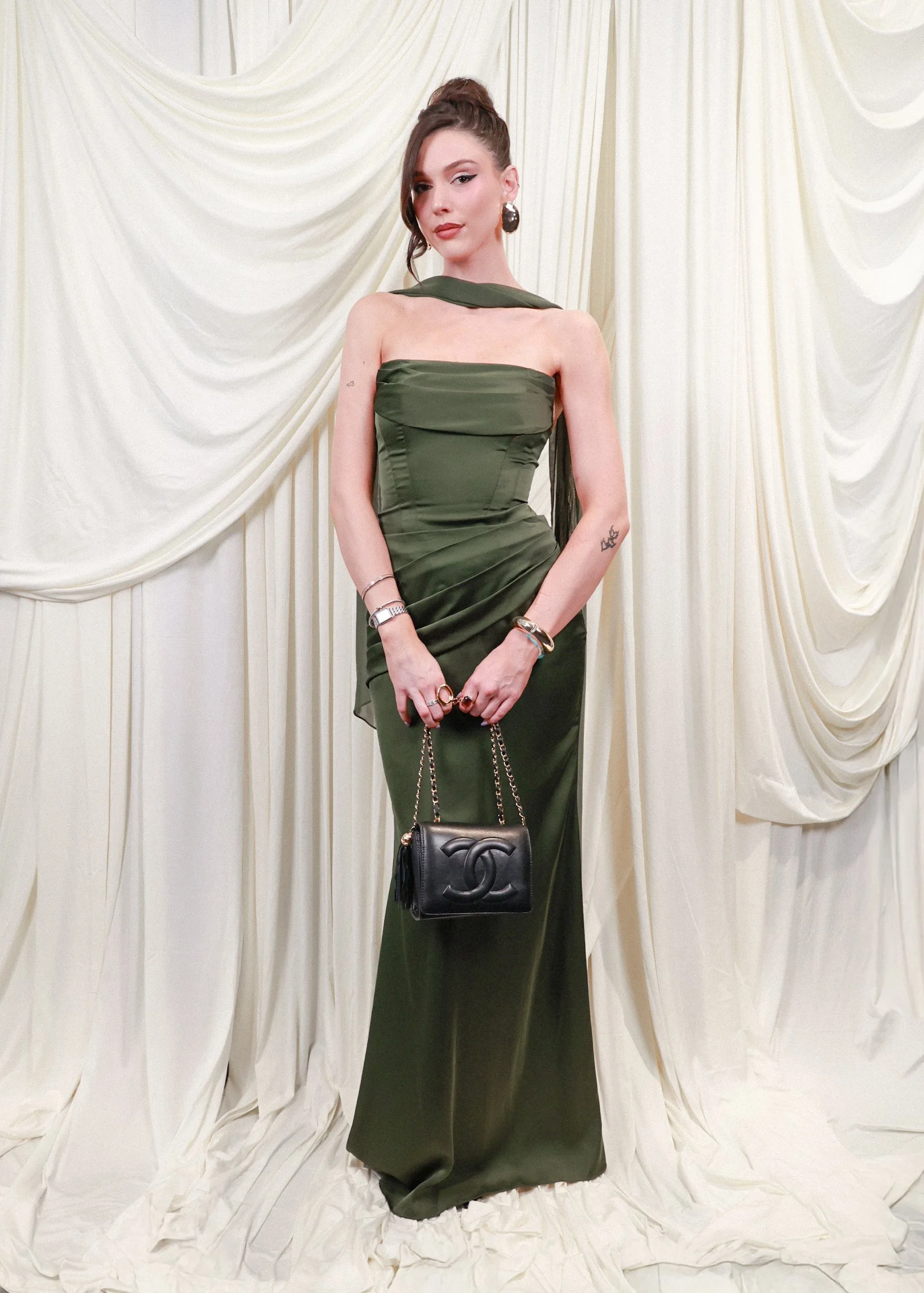 A woman with dark hair in an updo hairstyle, wearing a strapless green evening gown, accessorized with large earrings, bracelets, rings, and holding a black Chanel purse, standing against cream-colored draped fabric.