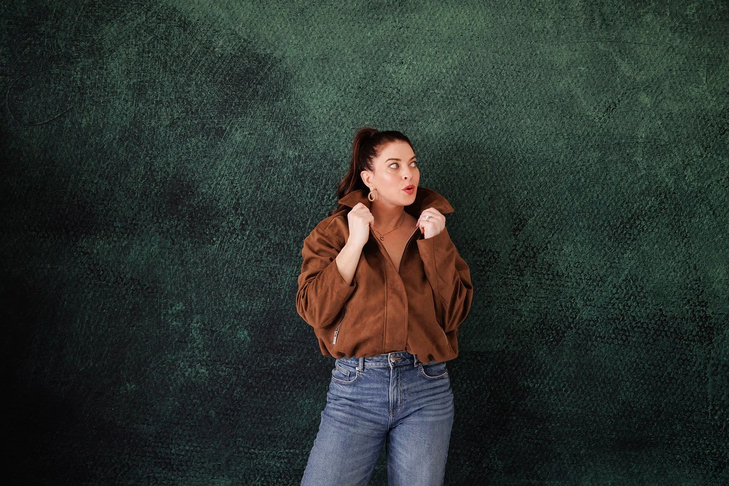 A woman with dark hair in a high ponytail wearing a brown jacket and blue jeans standing against a textured green background, looking to her left with an expressive facial expression.