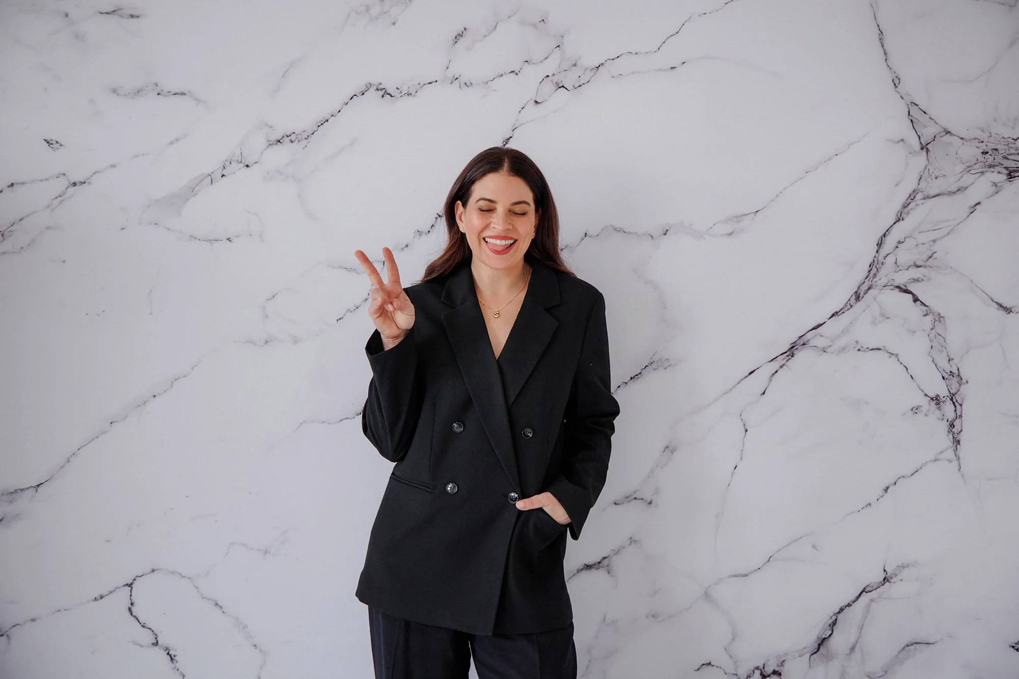 A smiling woman with medium-length dark hair making a peace sign with her right hand, standing against a white marble wall.