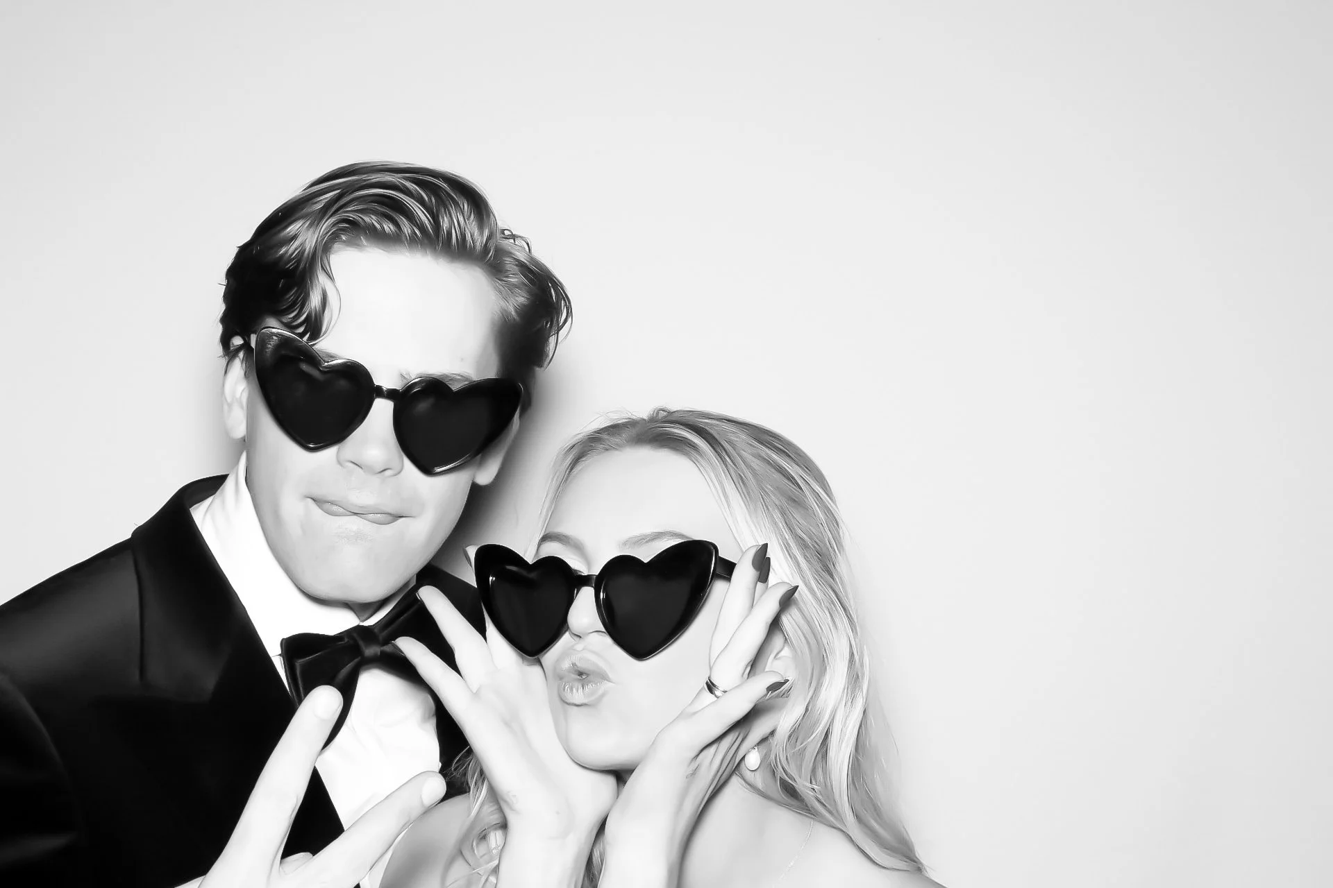 A black and white photo of a man and woman wearing oversized heart-shaped sunglasses, with the man in a tuxedo making a peace sign and the woman with her hands on her face making a playful expression.