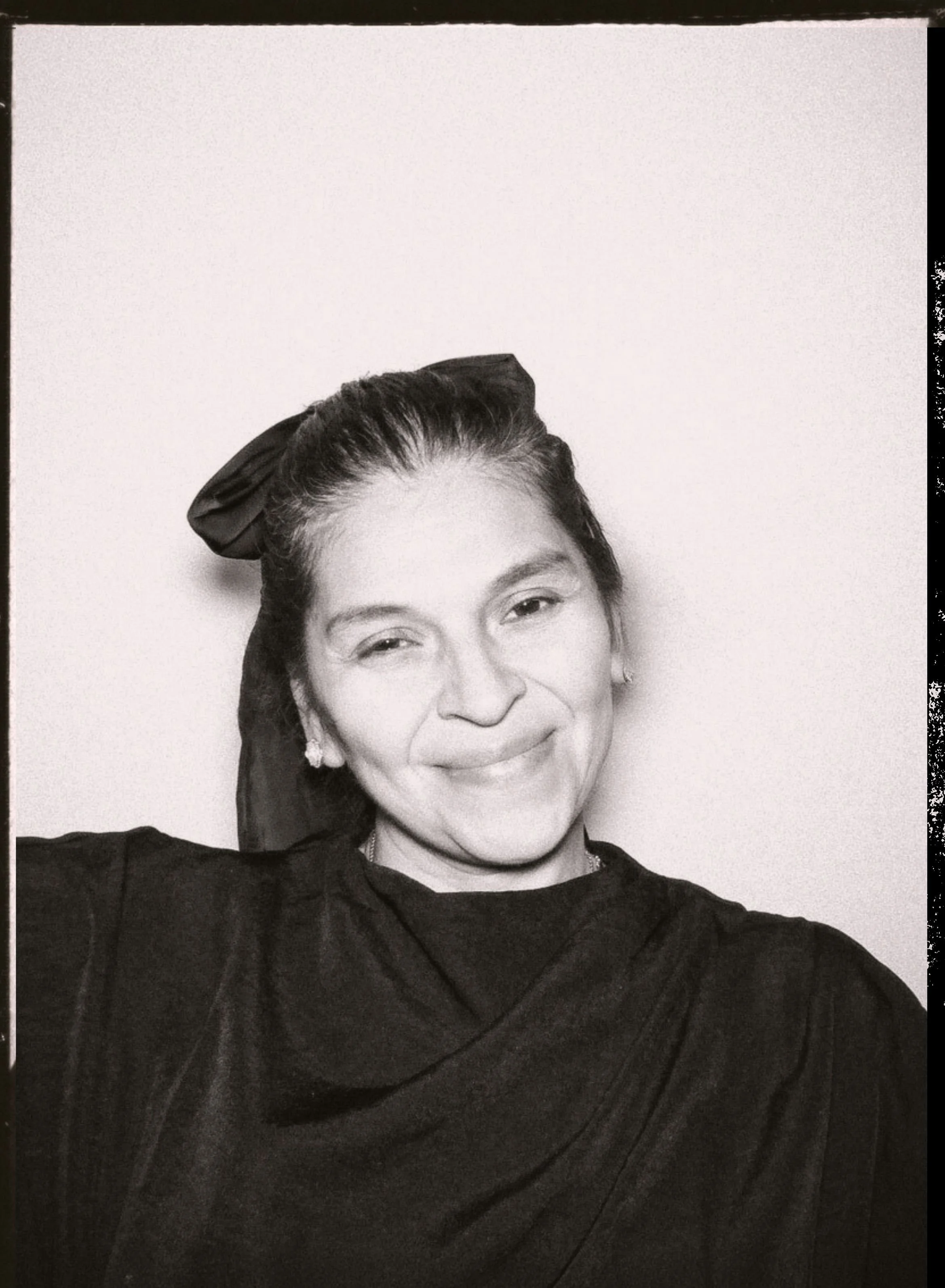 A woman taking a selfie in black and white, smiling with one eye closed, wearing a dark top and a large bow in her hair.