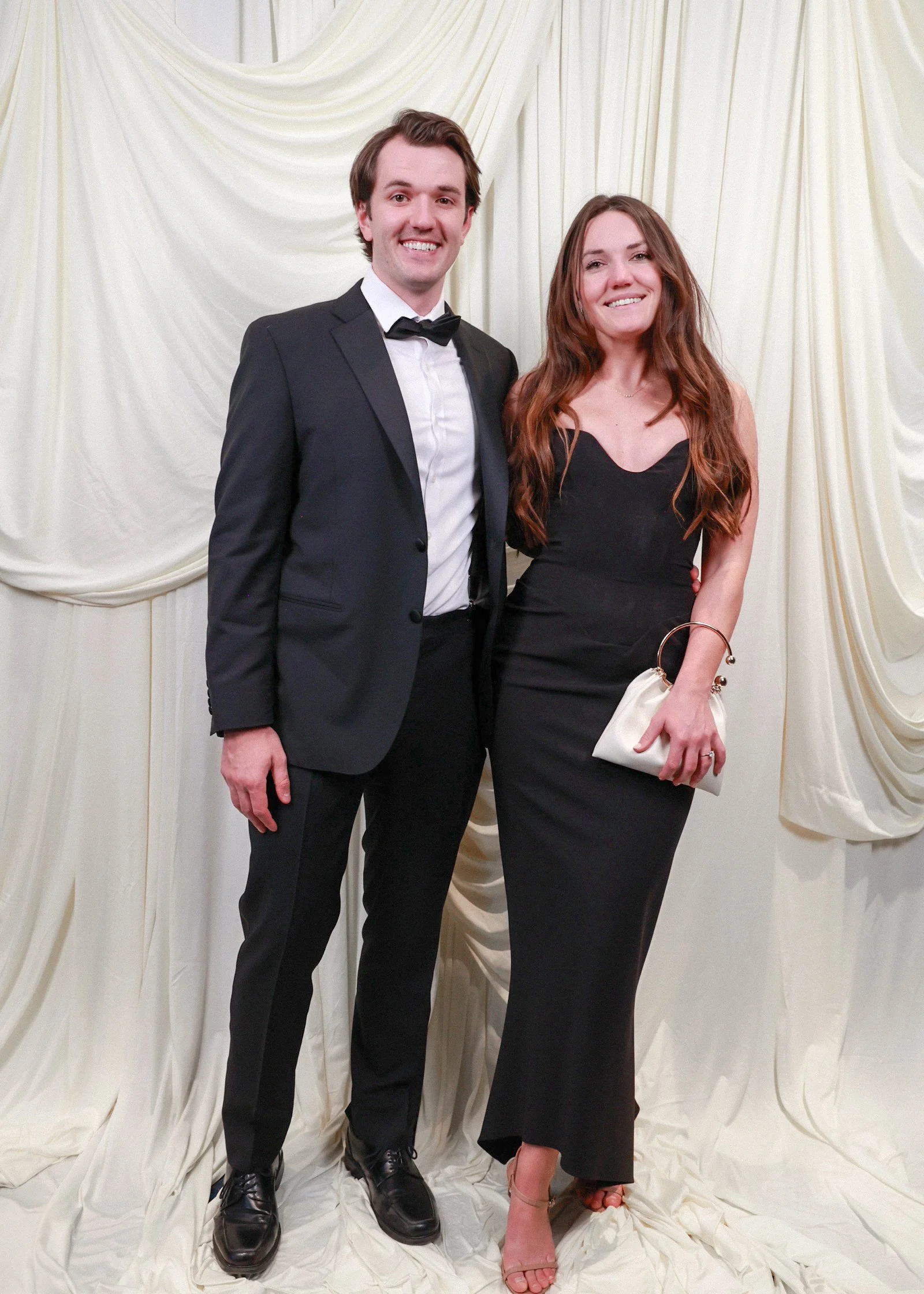 A man and woman standing together in front of a cream-colored draped backdrop, dressed formally. The man is wearing a black tuxedo with a white shirt and black bow tie, and the woman is wearing a fitted black dress, holding a small white clutch.