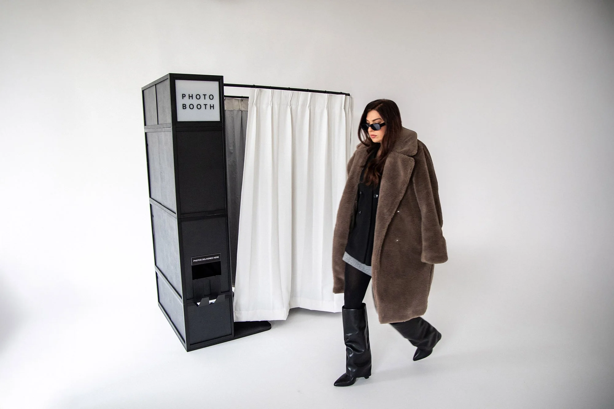 Woman in sunglasses and a brown fur coat walking past a photo booth with white curtains