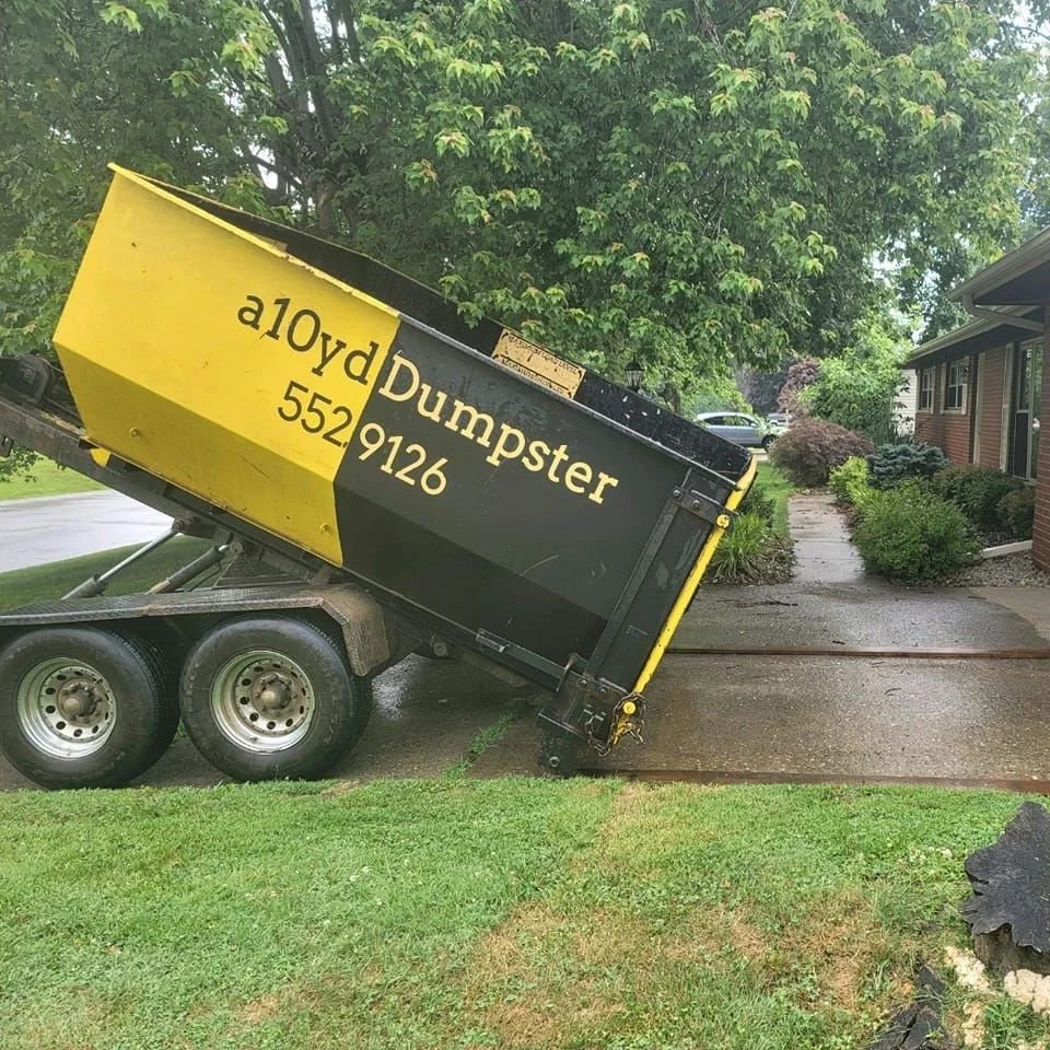 A dump trailer with yellow and black coloring, labeled 'a10yd Dumpster' and a phone number, tipped over on a driveway next to a lawn, with trees and a house in the background.