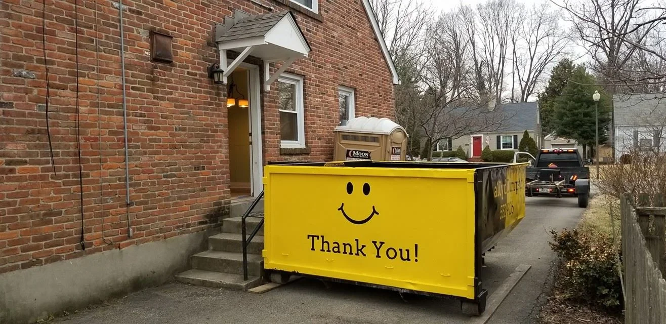 A yellow and black dumpster with a yellow front panel and a smiley face, parked outside a brick building with steps leading to the door, in a neighborhood with trees and cars nearby.