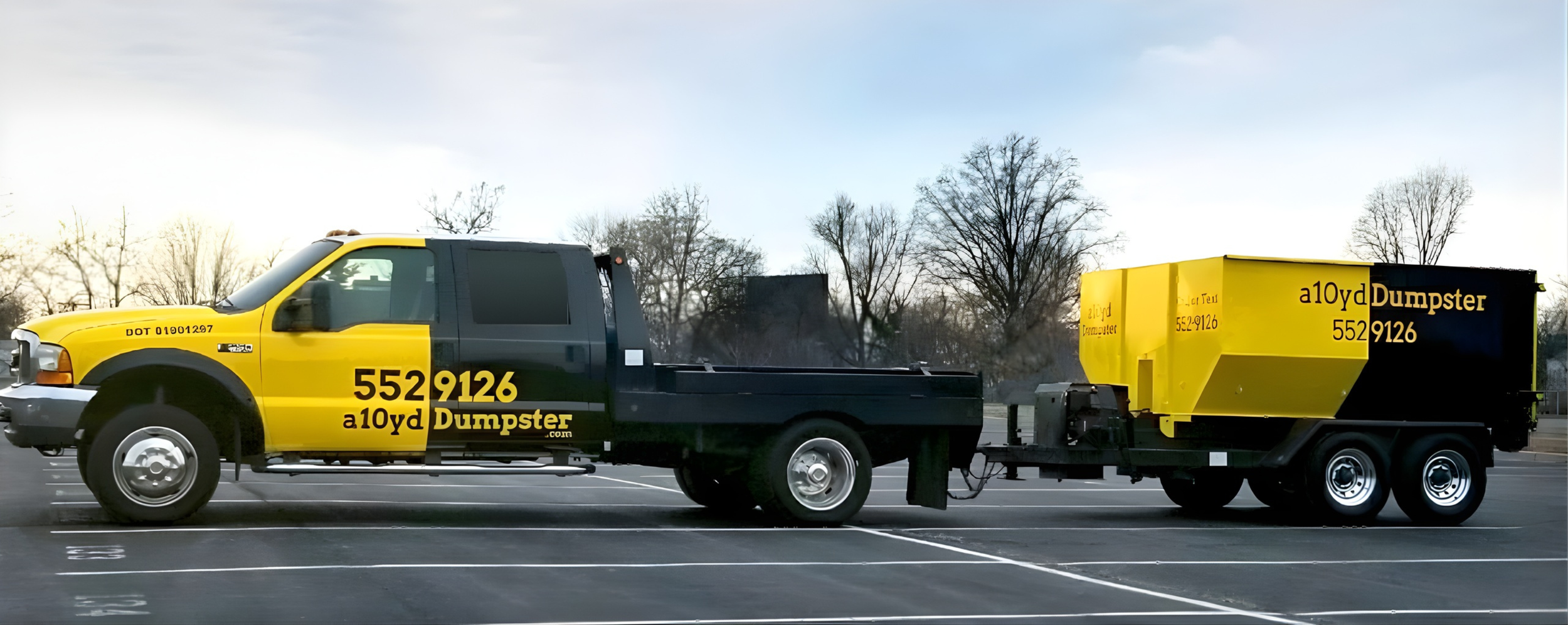 A yellow and black dumpster truck parked in a parking lot with trees in the background.
