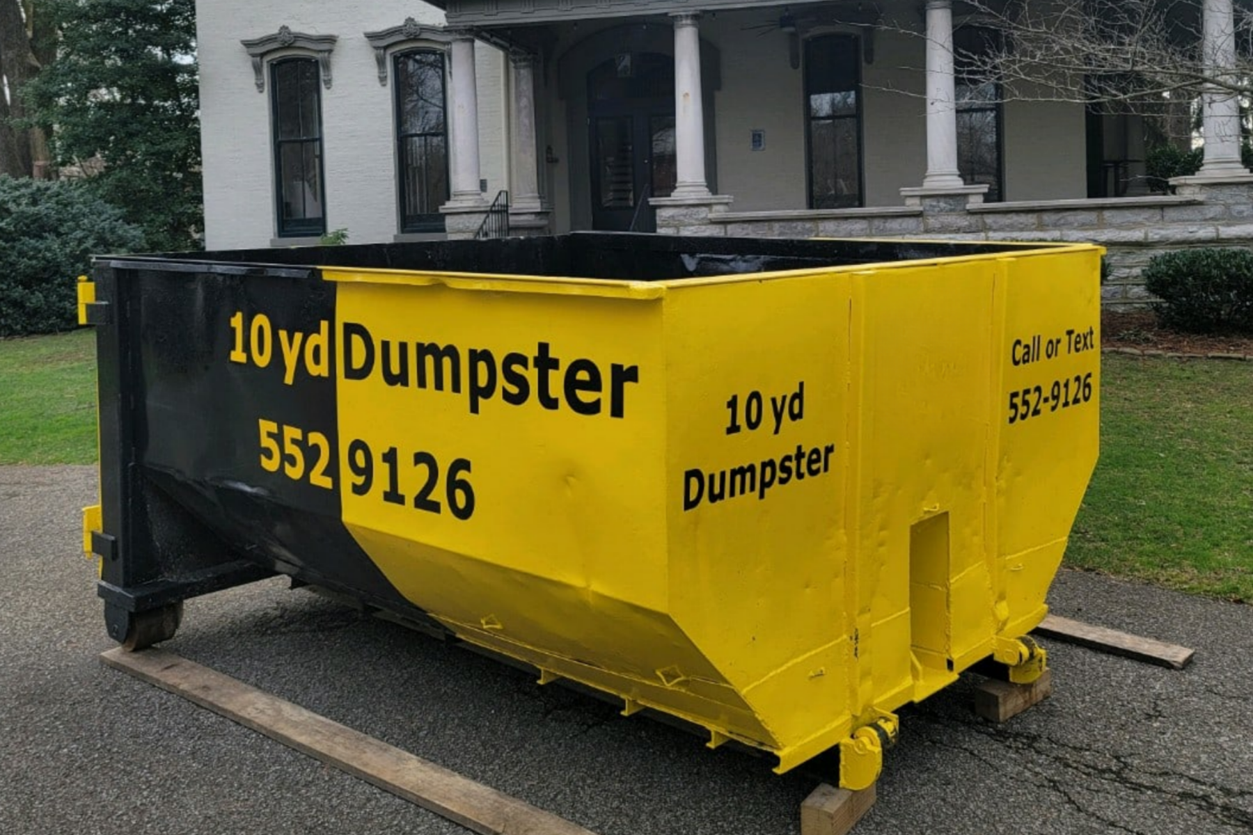 A large yellow and black dumpster labeled '10 yd Dumpster' with a phone number '552-9126' is placed on a driveway in front of a house with a porch, columns, and tall windows.