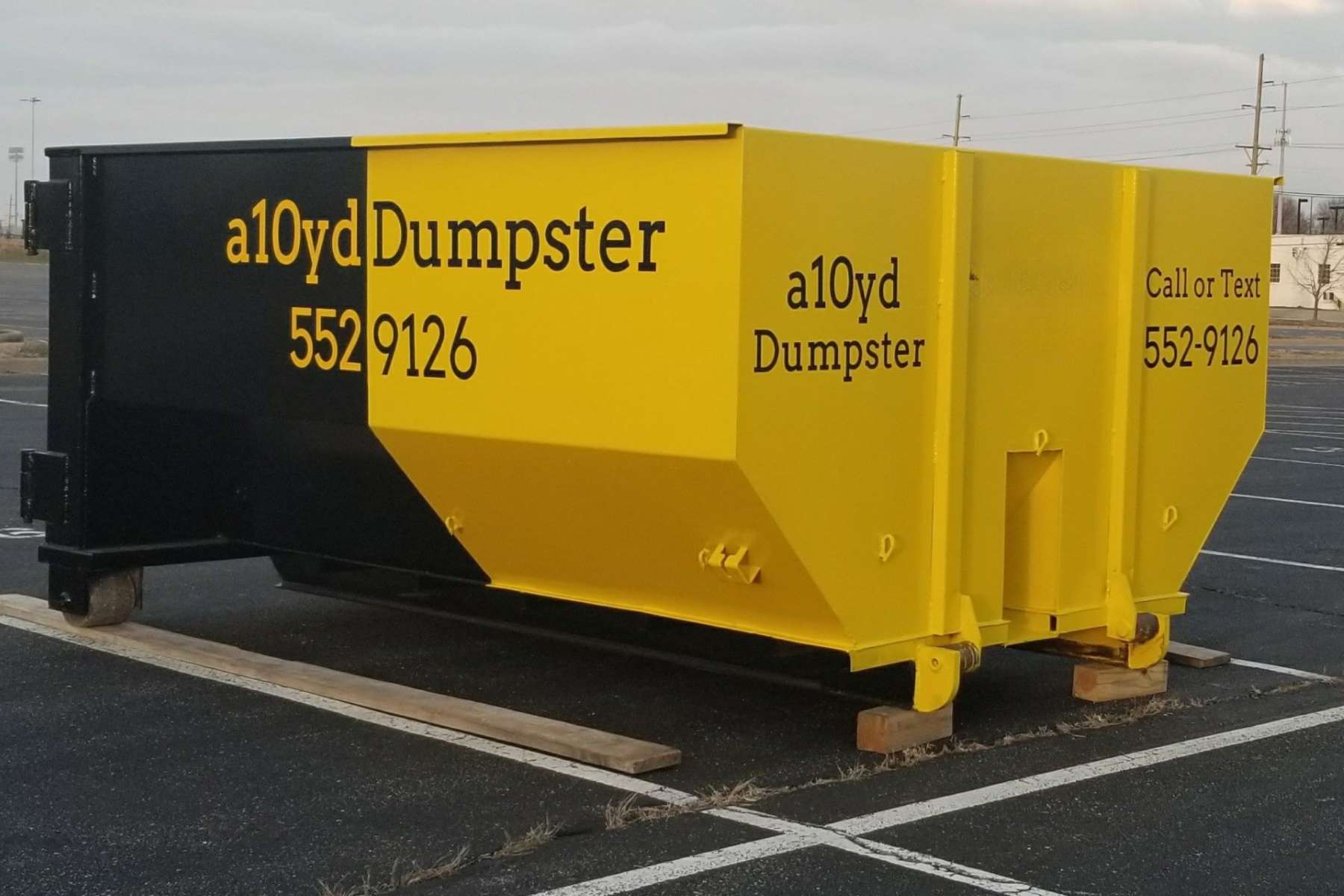 A large, yellow and black dumpster with the words 'a10yd Dumpster' and a phone number, placed in a parking lot.
