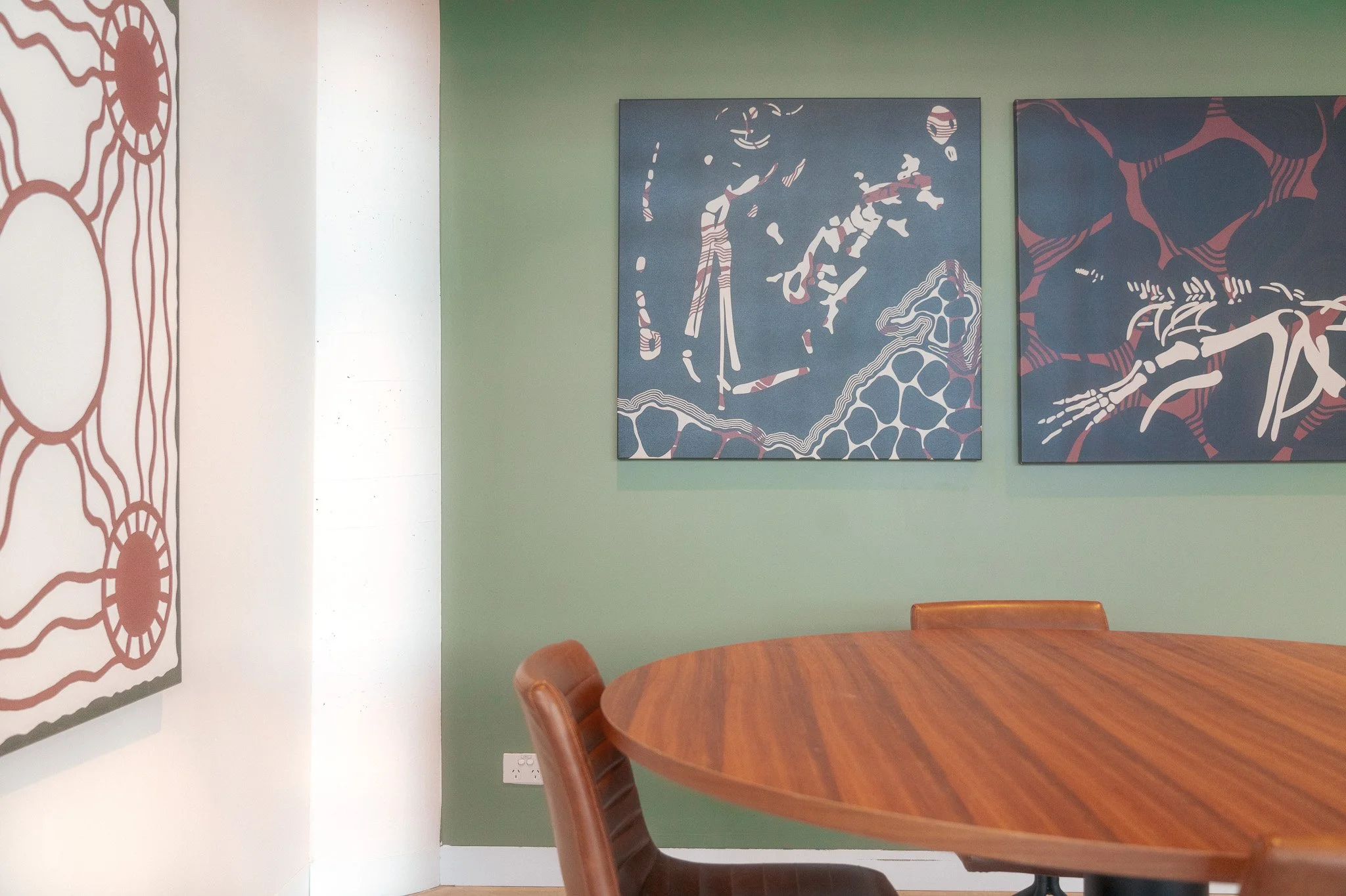 A dining table with a tray holding cups, a teapot, and desserts, against a green wall with two abstract artworks.