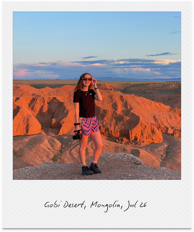 A woman standing on a rocky outcrop in Gobi Desert, Mongolia, during sunset, with a camera in one hand and smiling, wearing sunglasses, a black shirt, and colorful shorts.