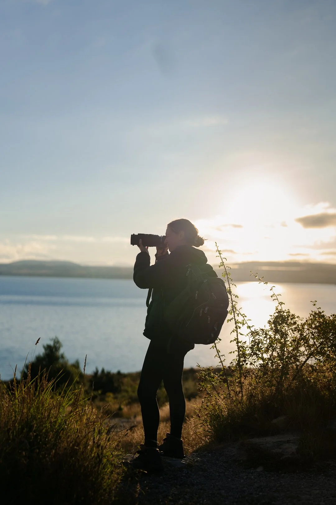 A person with a backpack standing on a trail near water, looking through binoculars at sunset.