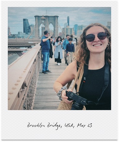 Smiling woman with sunglasses holding a camera on Brooklyn Bridge, with other pedestrians and Manhattan skyline in background.