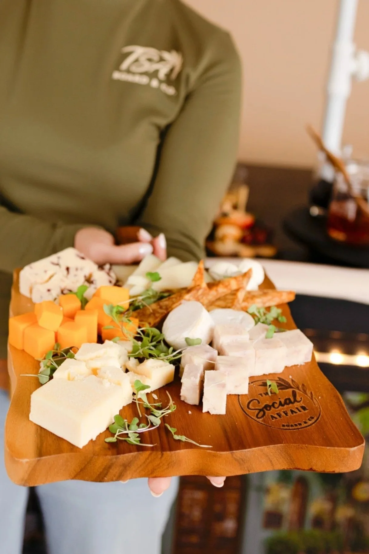 A person holding a wooden cheese board with assorted cheeses, carrots, and garnishes, with a person in a green shirt in the background.