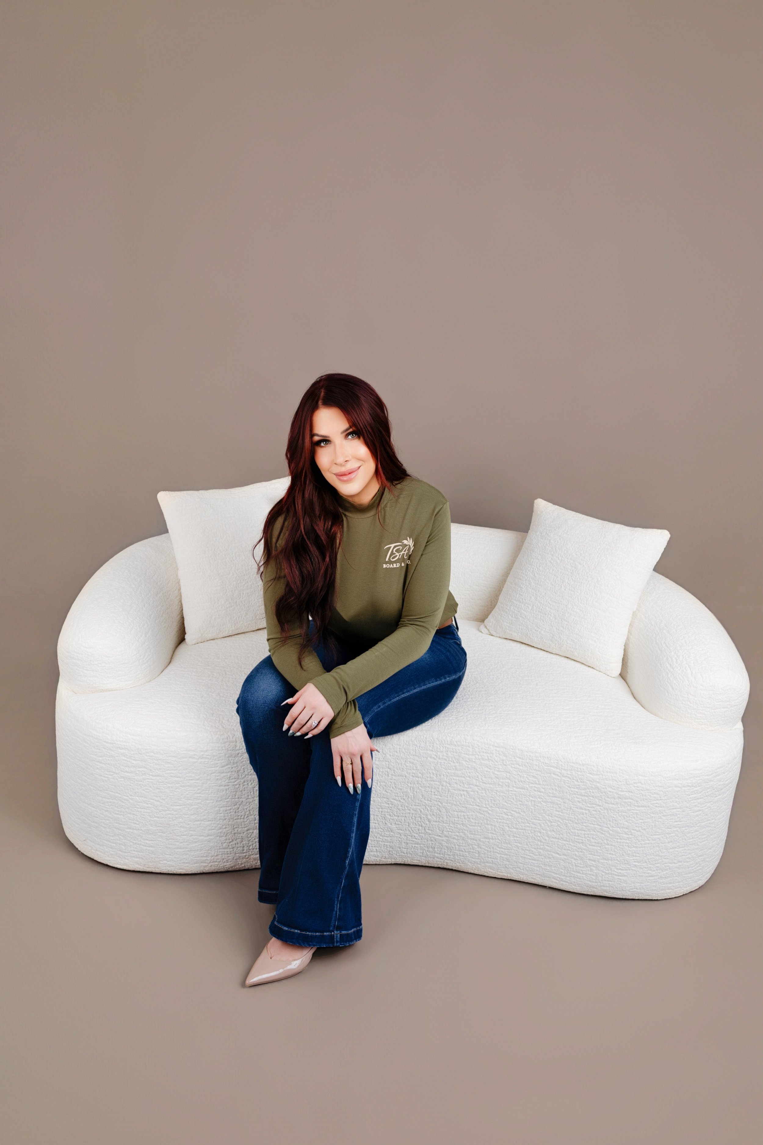 A woman with long dark hair and a green long-sleeve shirt sitting on a white, modern sofa with two white pillows against a plain beige background.