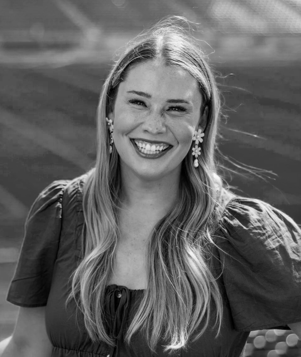 A woman with long hair, smiling, wearing earrings and a dress with puffed sleeves, outdoor setting.