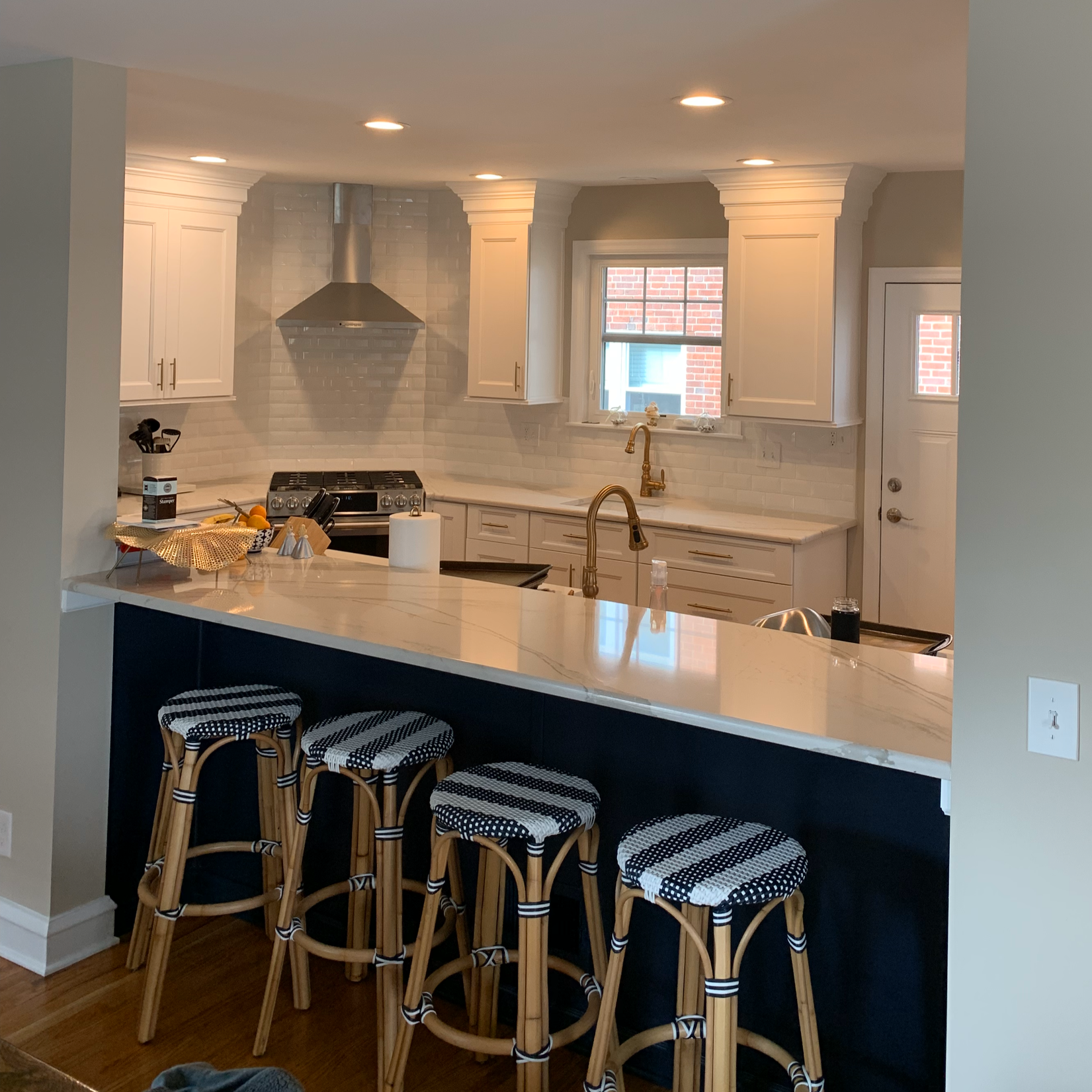 Modern kitchen with white cabinetry, a marble countertop breakfast bar with four rattan stools, a stainless steel stove, a gold faucet, white subway tile backsplash, a window with a view of a brick building, and recessed lighting.