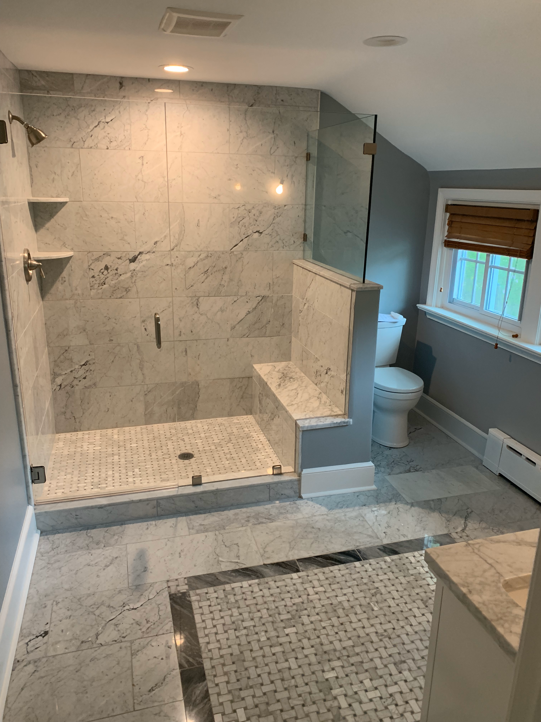 A modern bathroom with a marble walk-in shower, some small corner shelves, a partial glass door, a toilet, a window with wooden blinds, and a tiled floor with a woven-style rug.