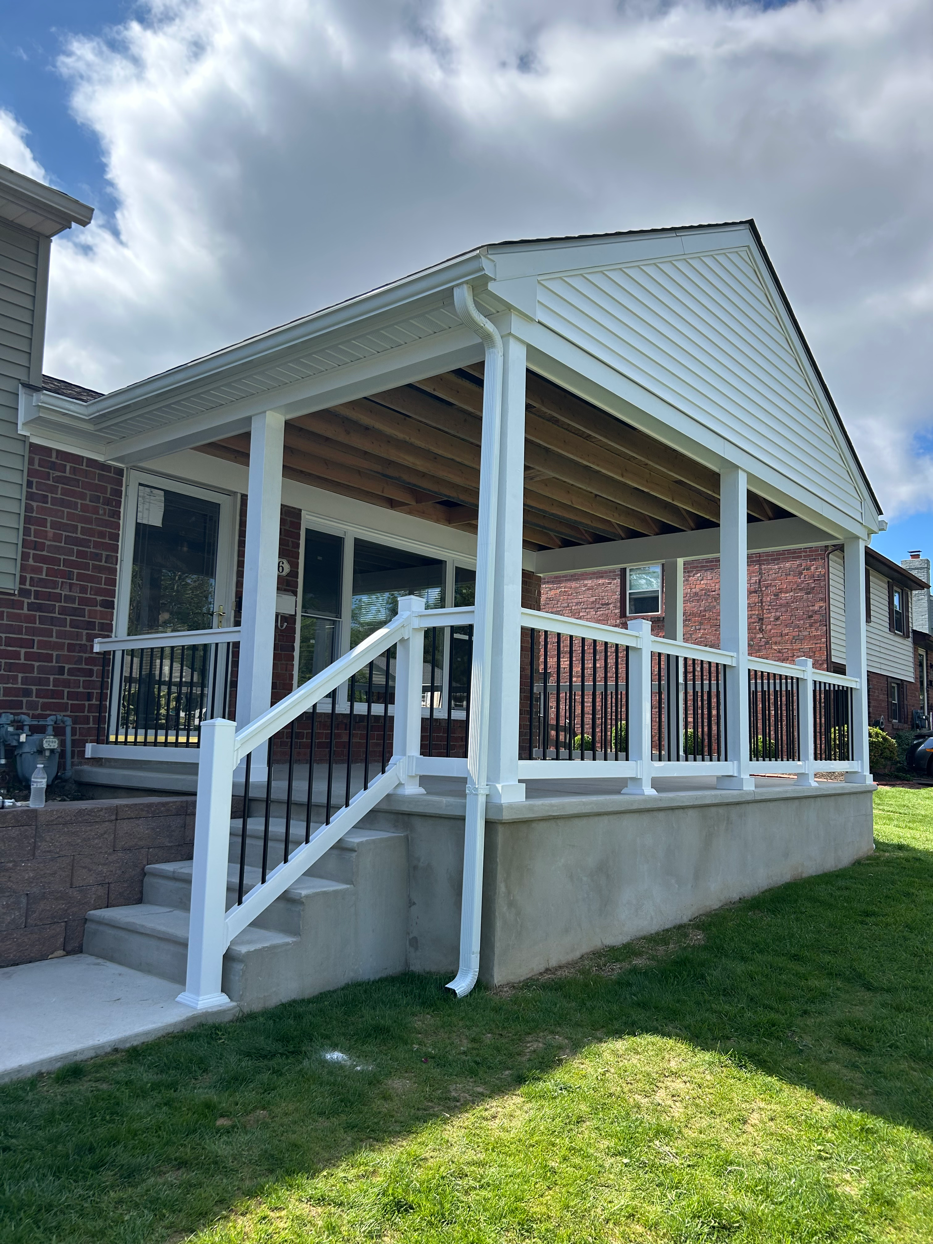 A house with a brick exterior and a raised porch with white railing and black balusters, accessed by concrete steps, under a blue sky with clouds.