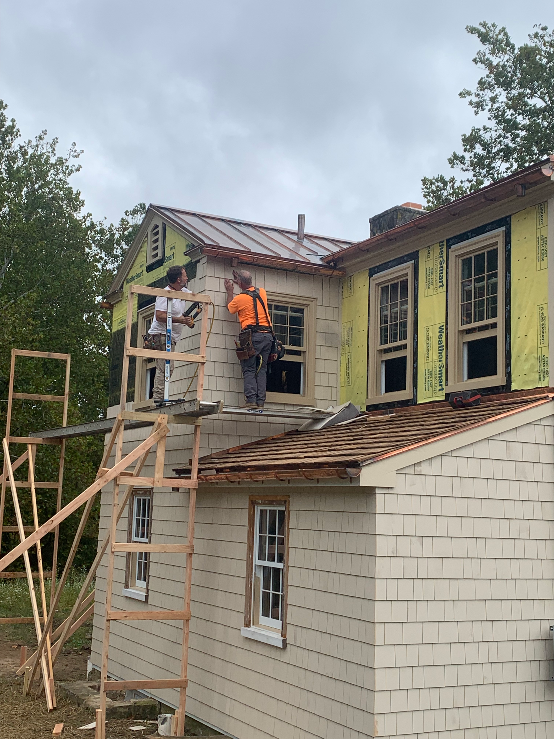 Two construction workers working on the exterior of a house, installing or repairing a window on the upper floor, with scaffolding and wooden framing around.
