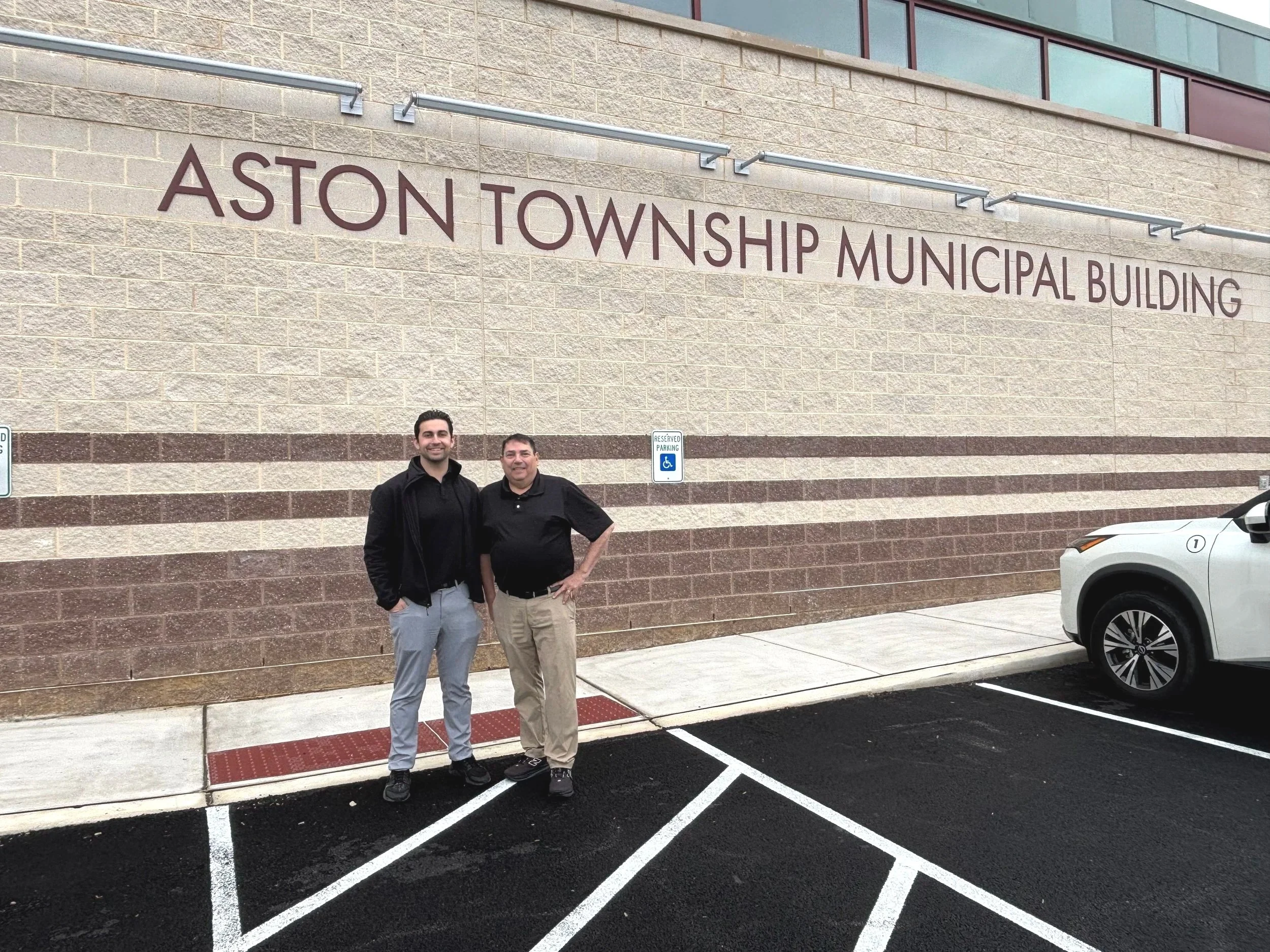 Two men standing side by side outside the Aston Township Municipal Building, smiling at the camera, in a parking lot with marked parking spaces and a reserved handicapped parking sign.
