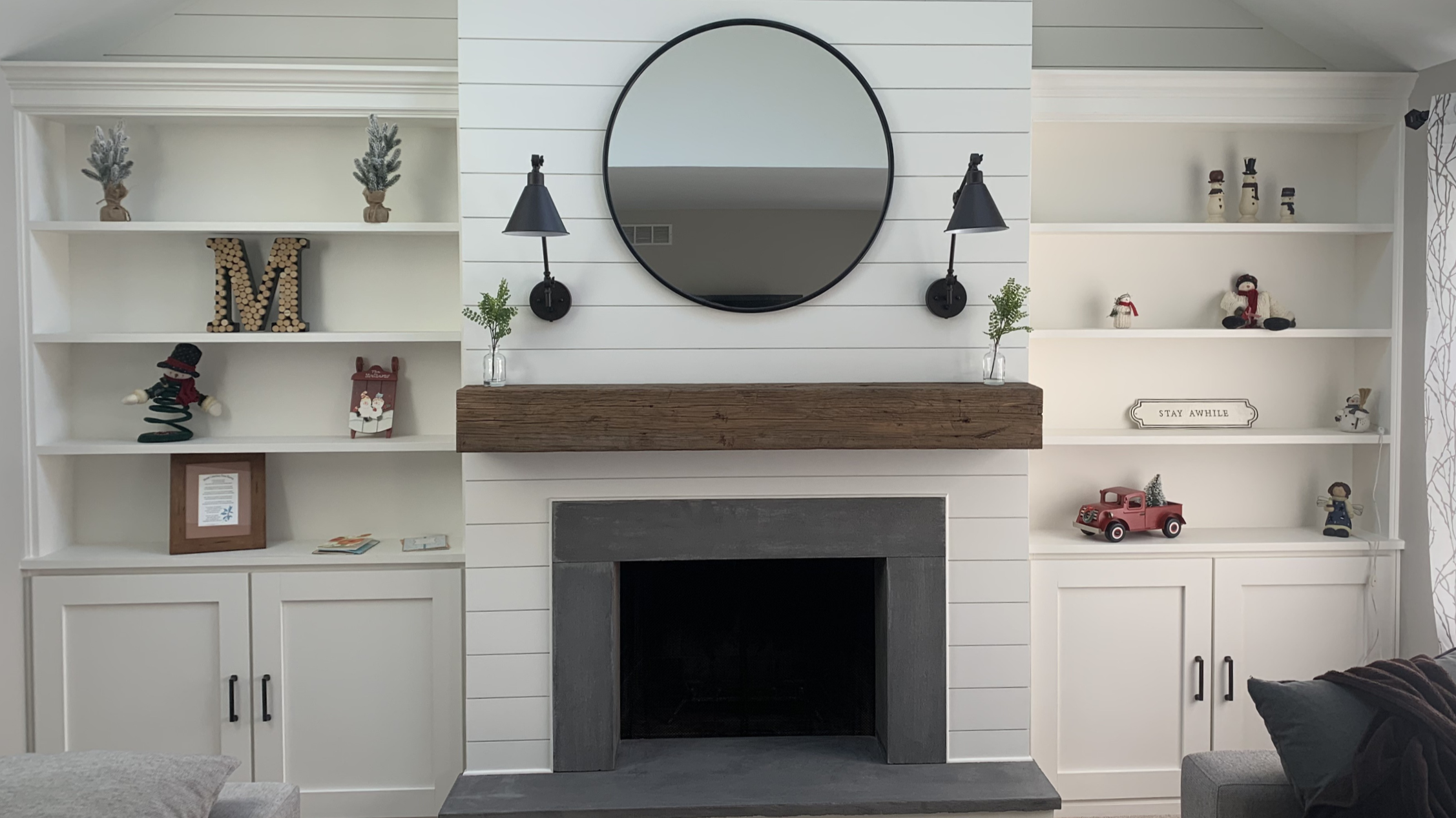 Living room with white built-in shelves decorated with Christmas figurines, a round mirror, black wall sconces, and a wooden mantel above a fireplace.