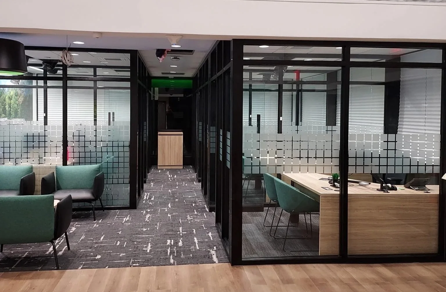 Modern office space with glass partition walls, green and black chairs, and a wooden desk with office supplies inside a glass-enclosed conference room.