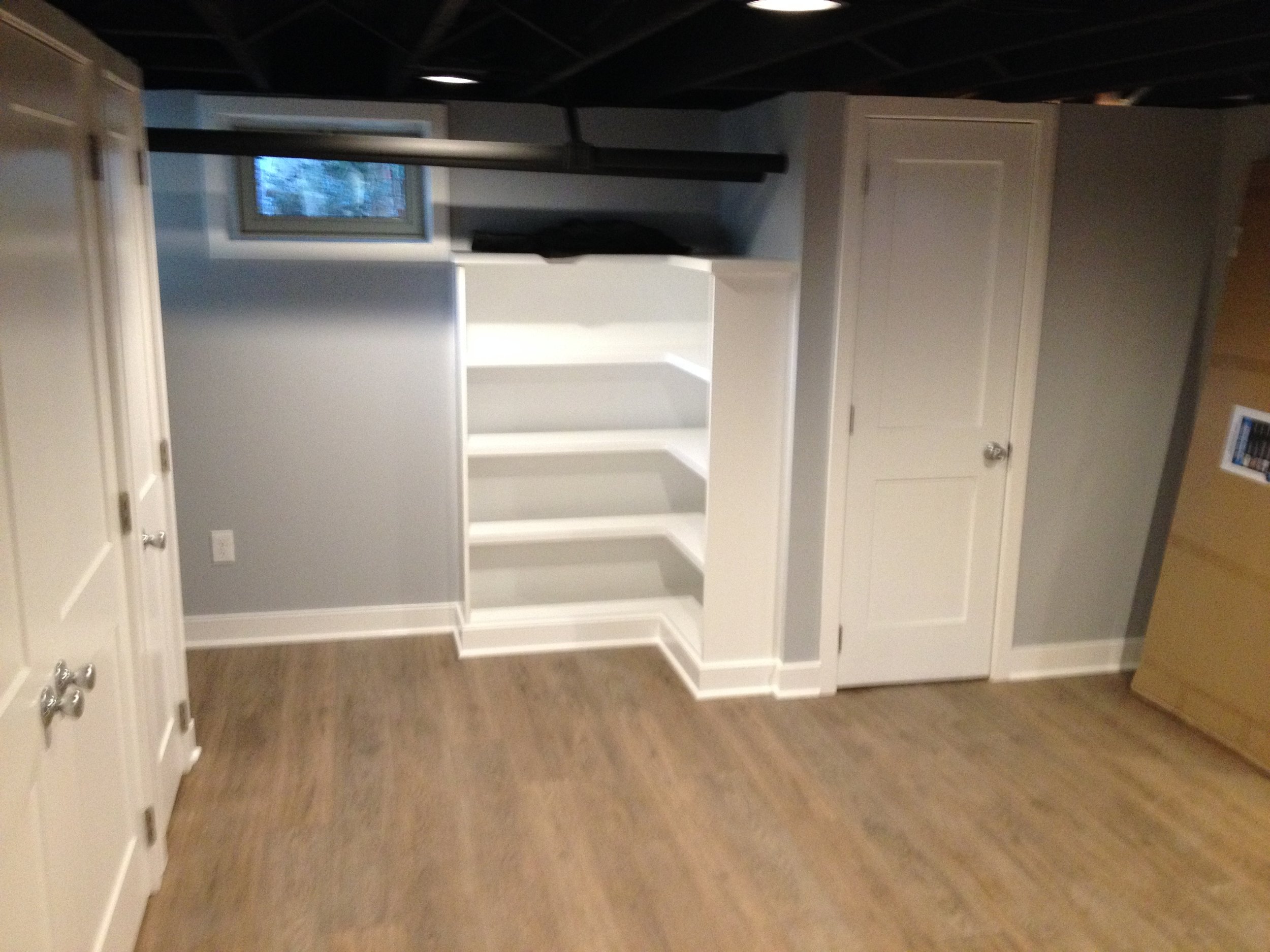 An empty room with hardwood floor, light gray walls, and white built-in shelves. There are two white doors on the right and a small window above the shelves. A black object is on top of the shelves.