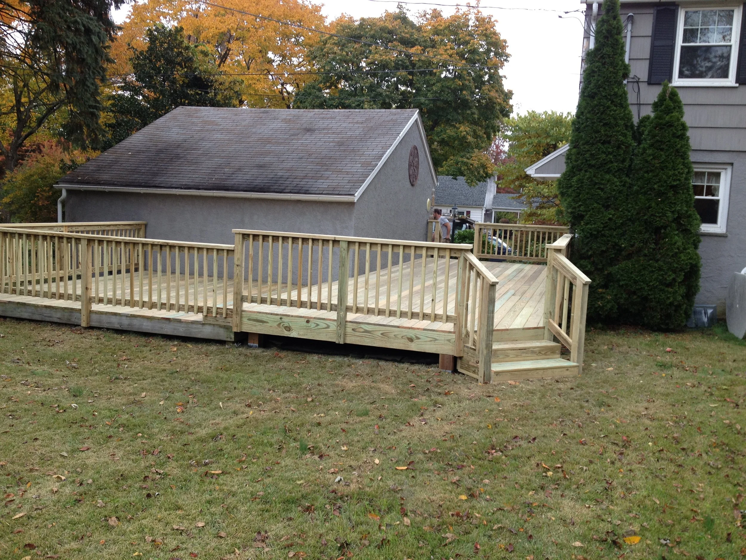 Newly built wooden deck with railings and stairs in a backyard, next to a house with grey siding, surrounded by trees with autumn leaves.