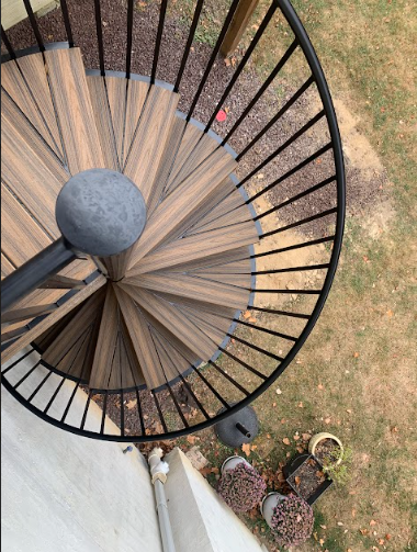 From above view of a spiral staircase with wooden steps and black metal railing, leading down to a grassy yard with potted plants and garden decorations.