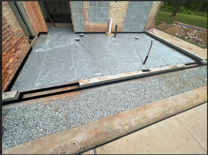 Construction site with a newly poured concrete slab outside a building, surrounded by gravel and a sidewalk.