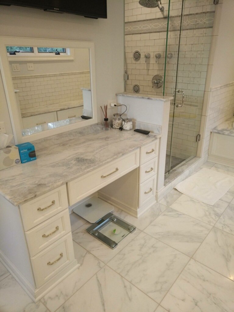 Bathroom with marble floor, vanity with marble countertop, mirror, and a glass-enclosed shower with multiple shower fixtures. There is a scale on the floor and some toiletries on the vanity.