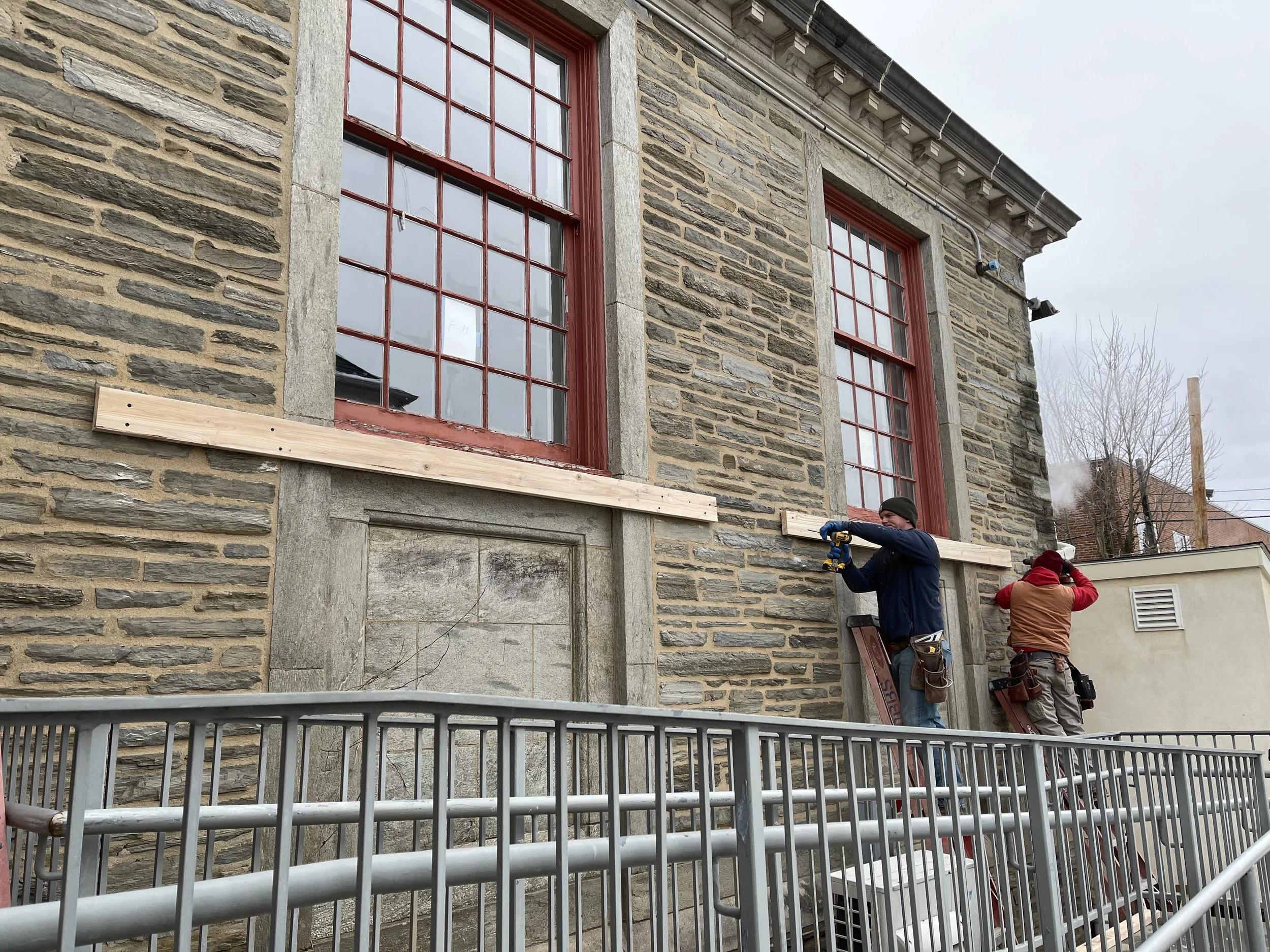 Two workers on the exterior stone wall of a building with large red-framed windows.