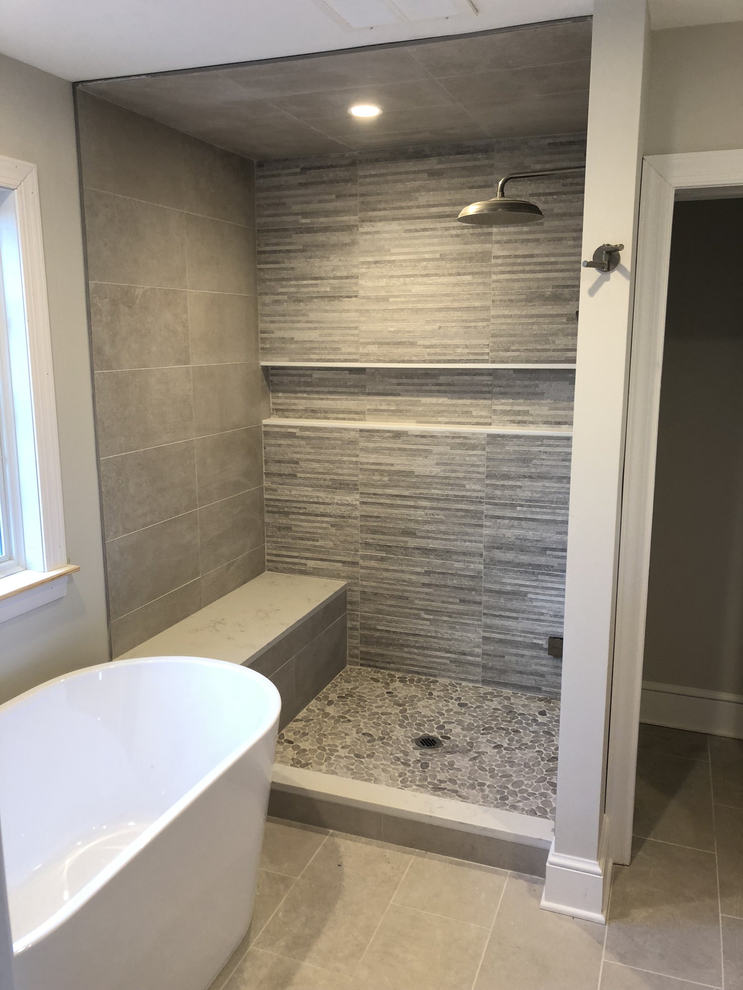 Modern walk-in shower with a pebble floor, gray tiled walls, a built-in bench, and a rain showerhead. A white bathtub is in the foreground, and there is a window with a white frame on the left side.