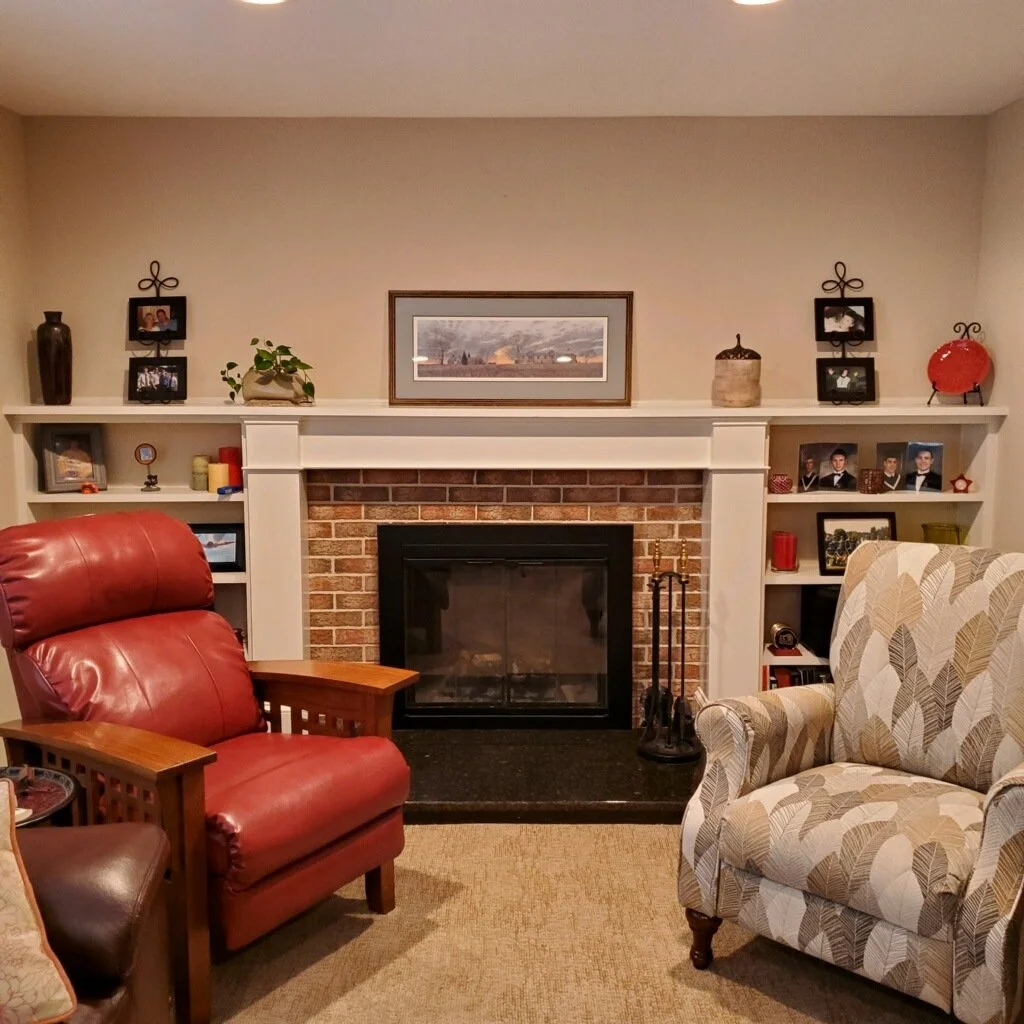 Living room with brick fireplace, surrounded by white built-in shelves and decorated with family photos, potted plant, vases, and artwork; red leather armchair and patterned upholstered armchair positioned near fireplace on carpeted floor.