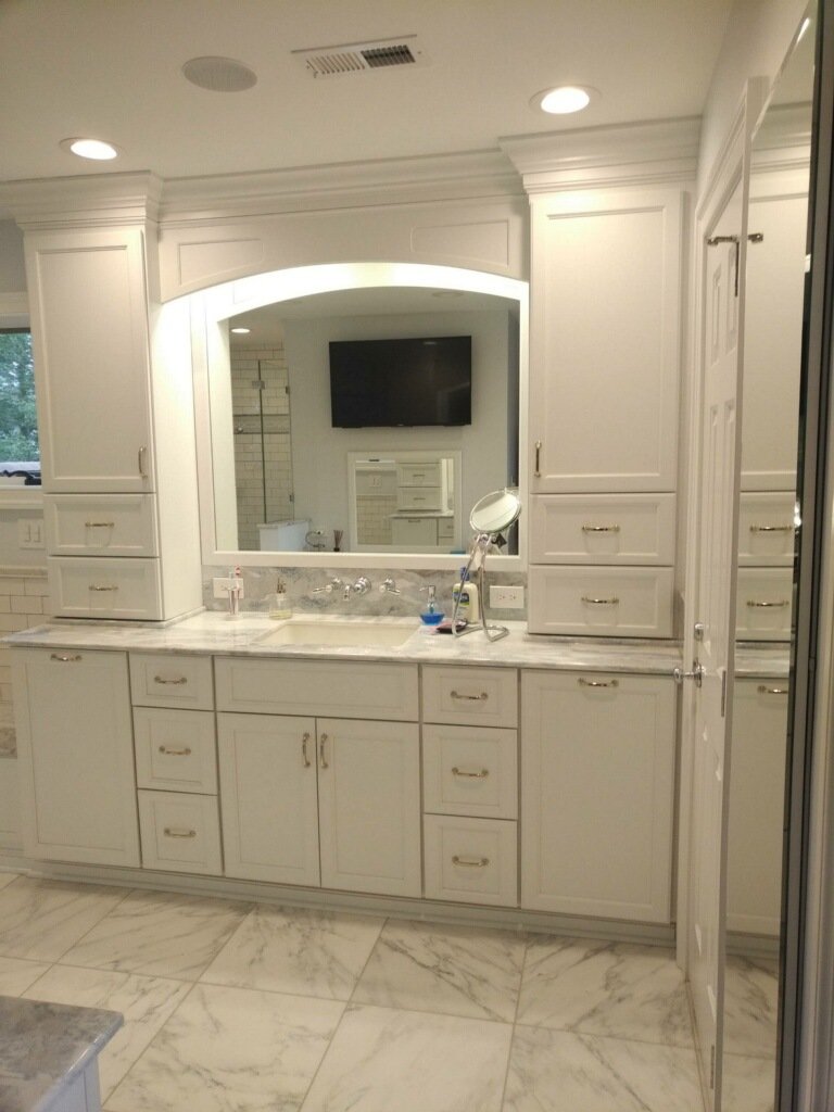 White bathroom vanity with a large mirror, marble countertop, and multiple drawers and cabinets. A small mirror and some toiletries are on the countertop. The floor is tiled with marble-look tiles, and a TV is mounted on the wall in the background.