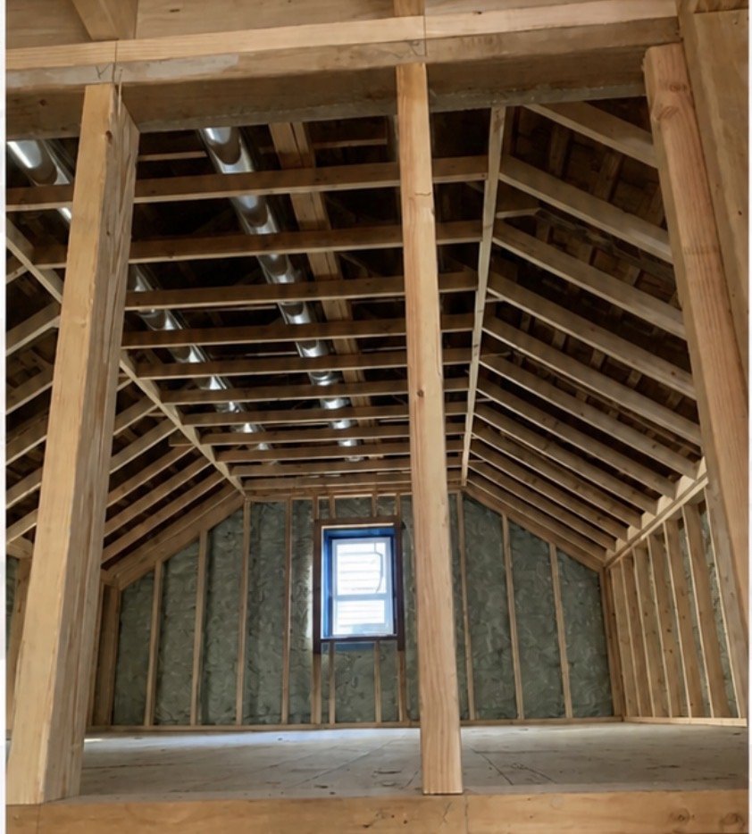 Interior of an unfinished attic with exposed wooden framing, roof sheathing, a small window, and visible ductwork in the ceiling.