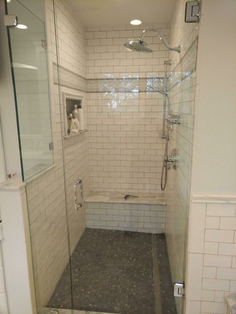 A walk-in shower with white subway tiles, a built-in shower seat, two showerheads, and a glass door, in a modern bathroom.