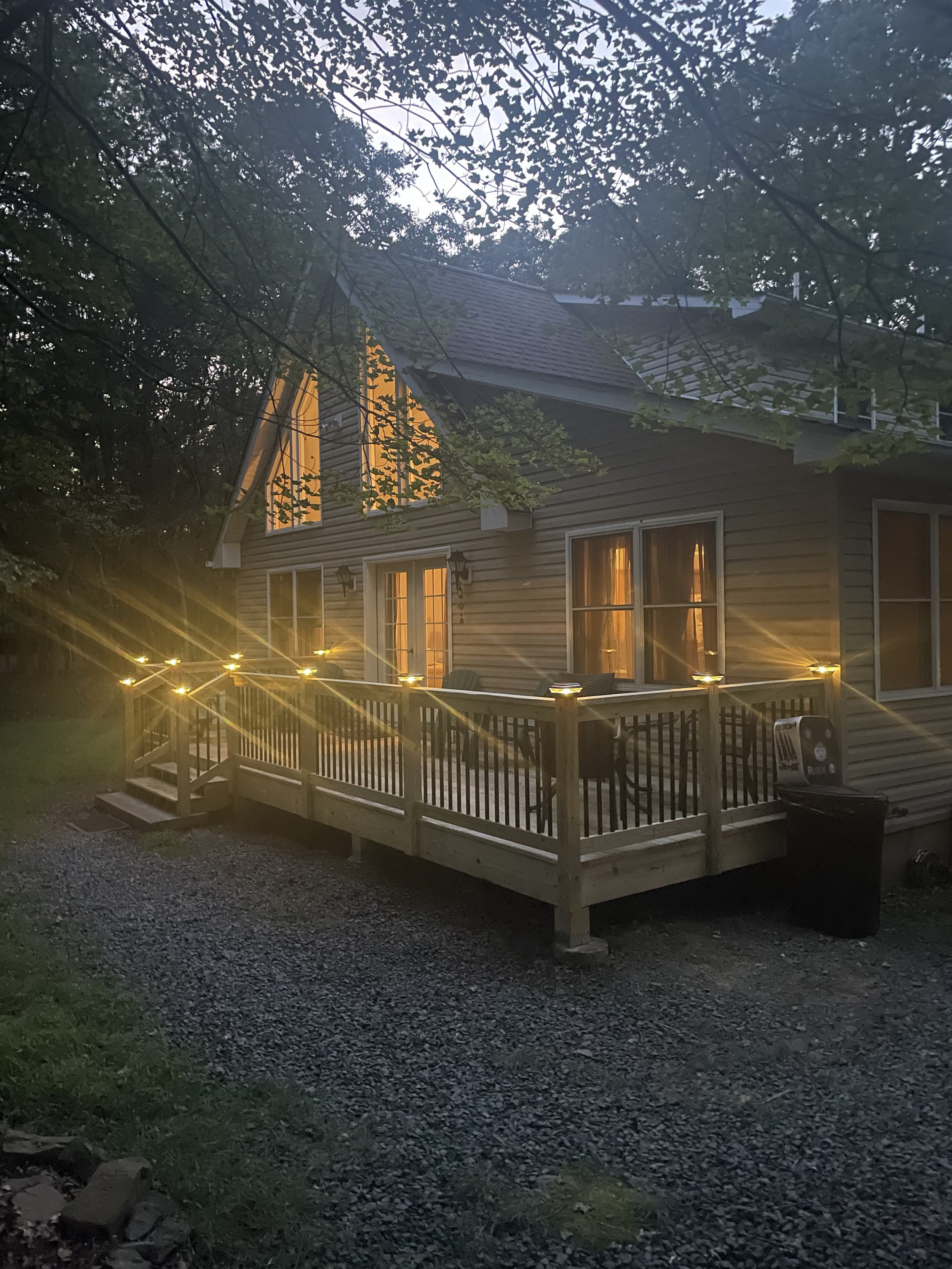 A house with a lit wooden deck and string lights, surrounded by trees at dusk.