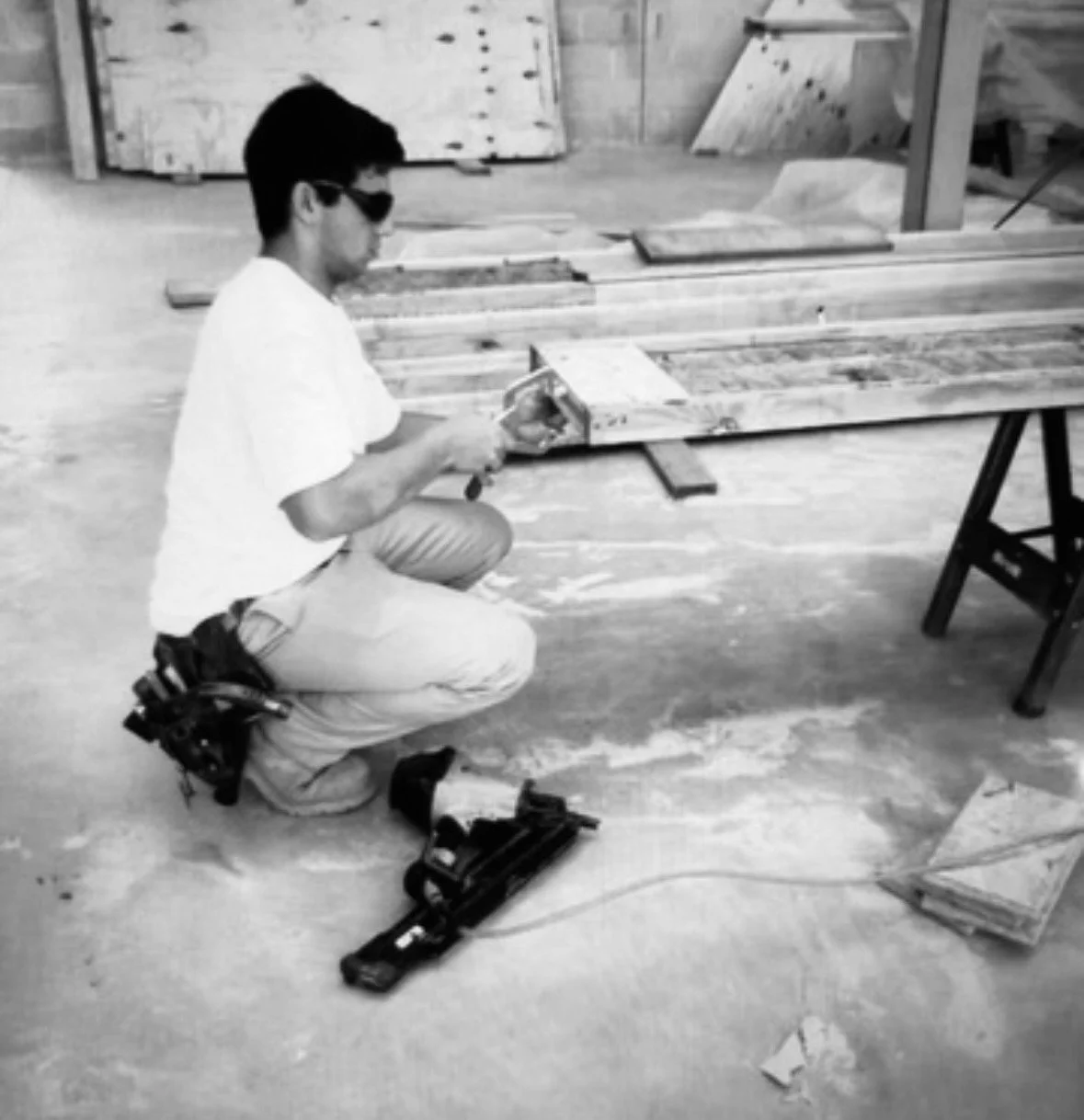 A man wearing sunglasses kneeling on the floor, using a hand saw on a piece of wood in a carpentry workshop.