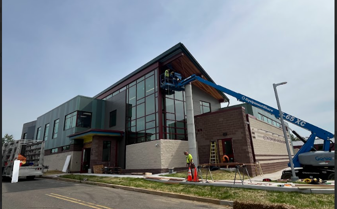 Construction workers installing large glass windows on a modern, multi-story building with a combination of metal and brick exterior, using a cherry picker and a ladder, on a street with equipment and safety cones.