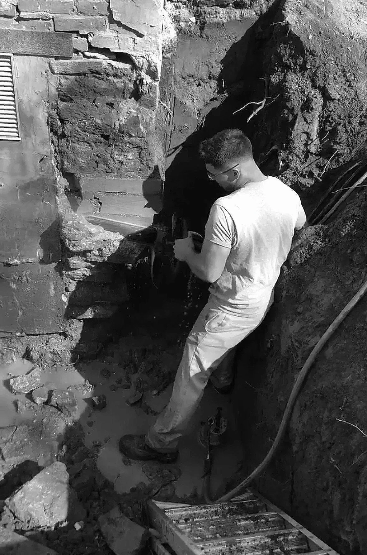 A man wearing glasses and a white t-shirt, holding a tool, stands in a construction site or excavation with a roughly dug trench in the ground. The trench is next to a building wall with brick and concrete. The man appears to be inspecting or working on the area, with construction materials and tools around.