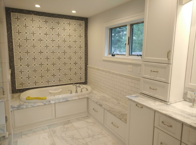Bathroom with a corner bathtub, white cabinets, marble countertops, a window, and tiled walls with a decorative wall feature.