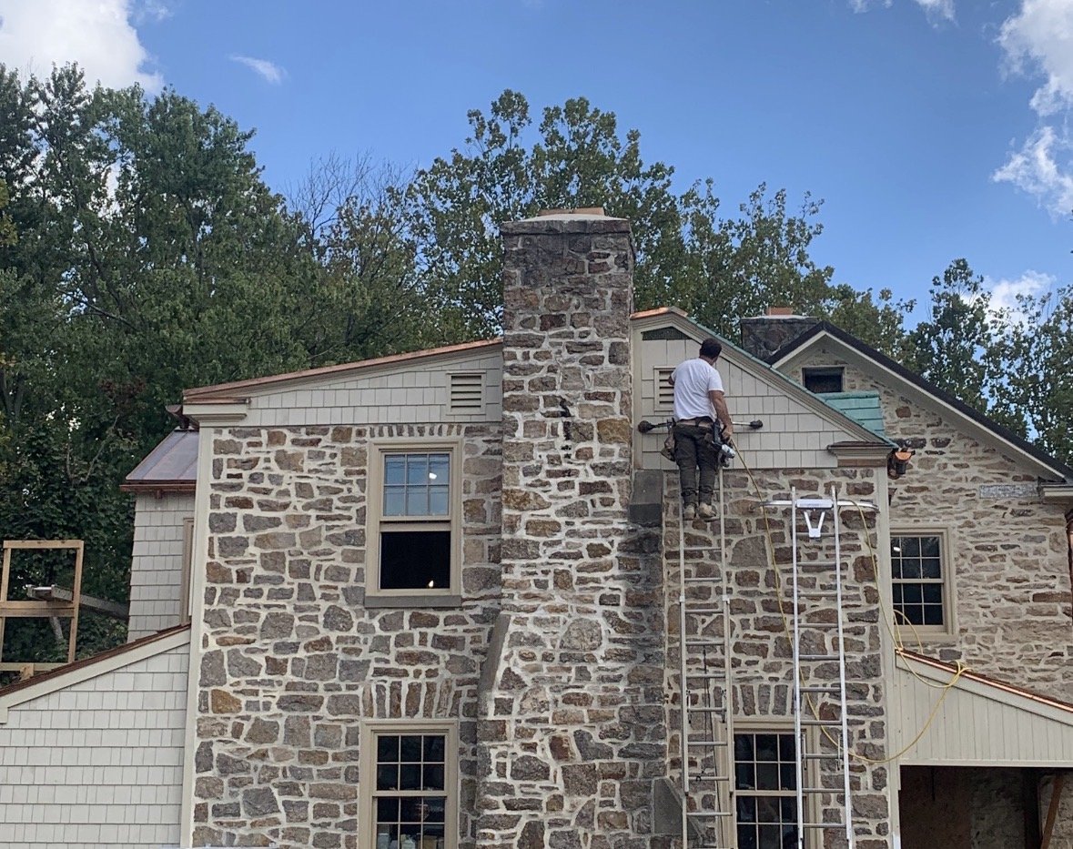 A worker repairing or inspecting a chimney on a stone and siding house, using scaffolding and tools, with a background of trees and blue sky.