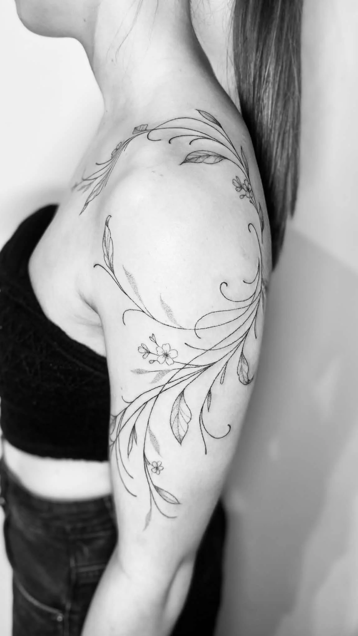 A black and white photo of a woman's upper arm, shoulder, and part of her face, with a floral tattoo featuring leaves and flowers on her shoulder.