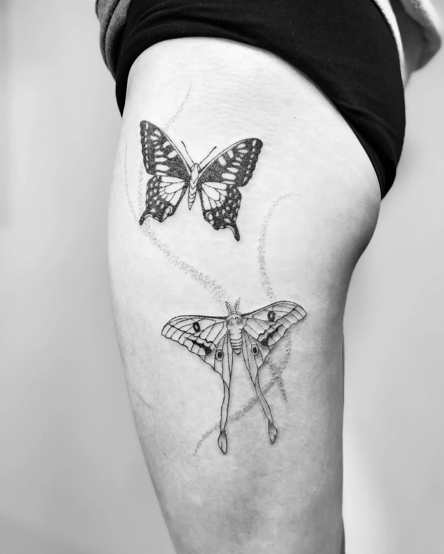 Black and white tattoo of two butterflies on a person's thigh, one above the other.